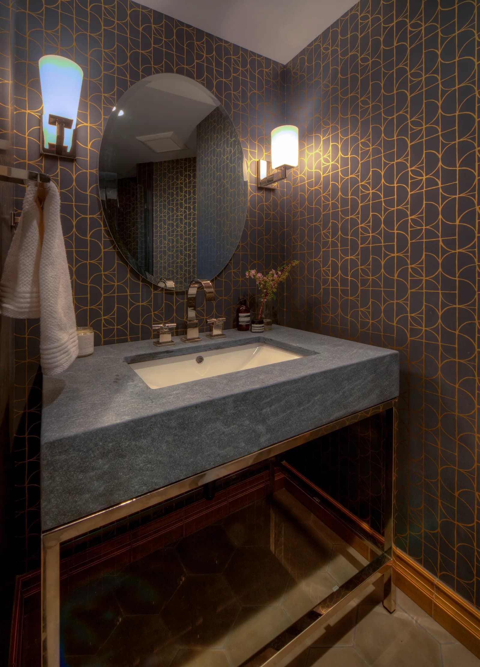 Powder room with gray and gold geometric wallpaper, an oval mirror, sconces, and a gray stone vanity on a polished metal base.