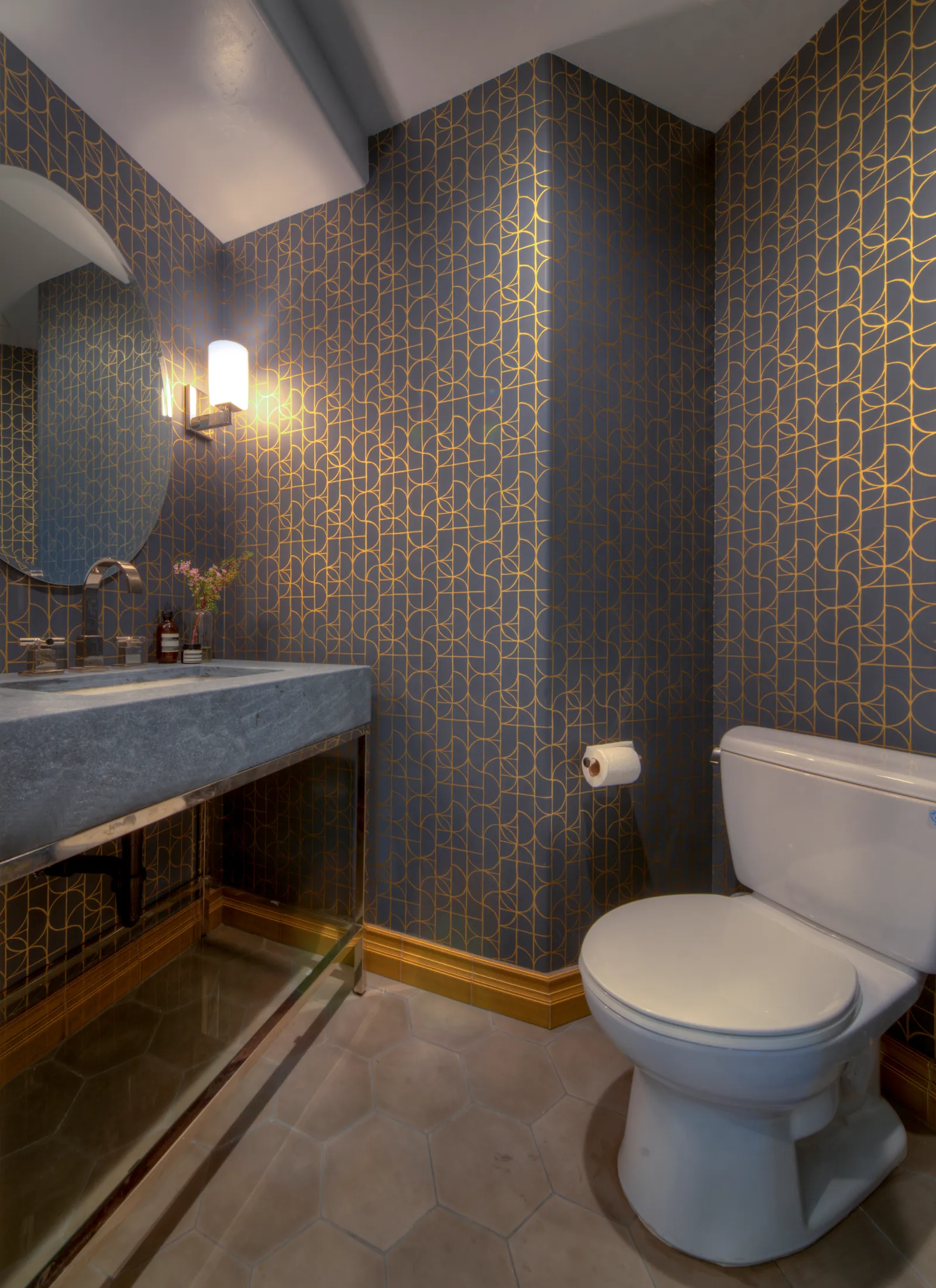 A powder room with navy and gold patterned tile walls, a gray stone trough sink, and hexagonal floor tile.