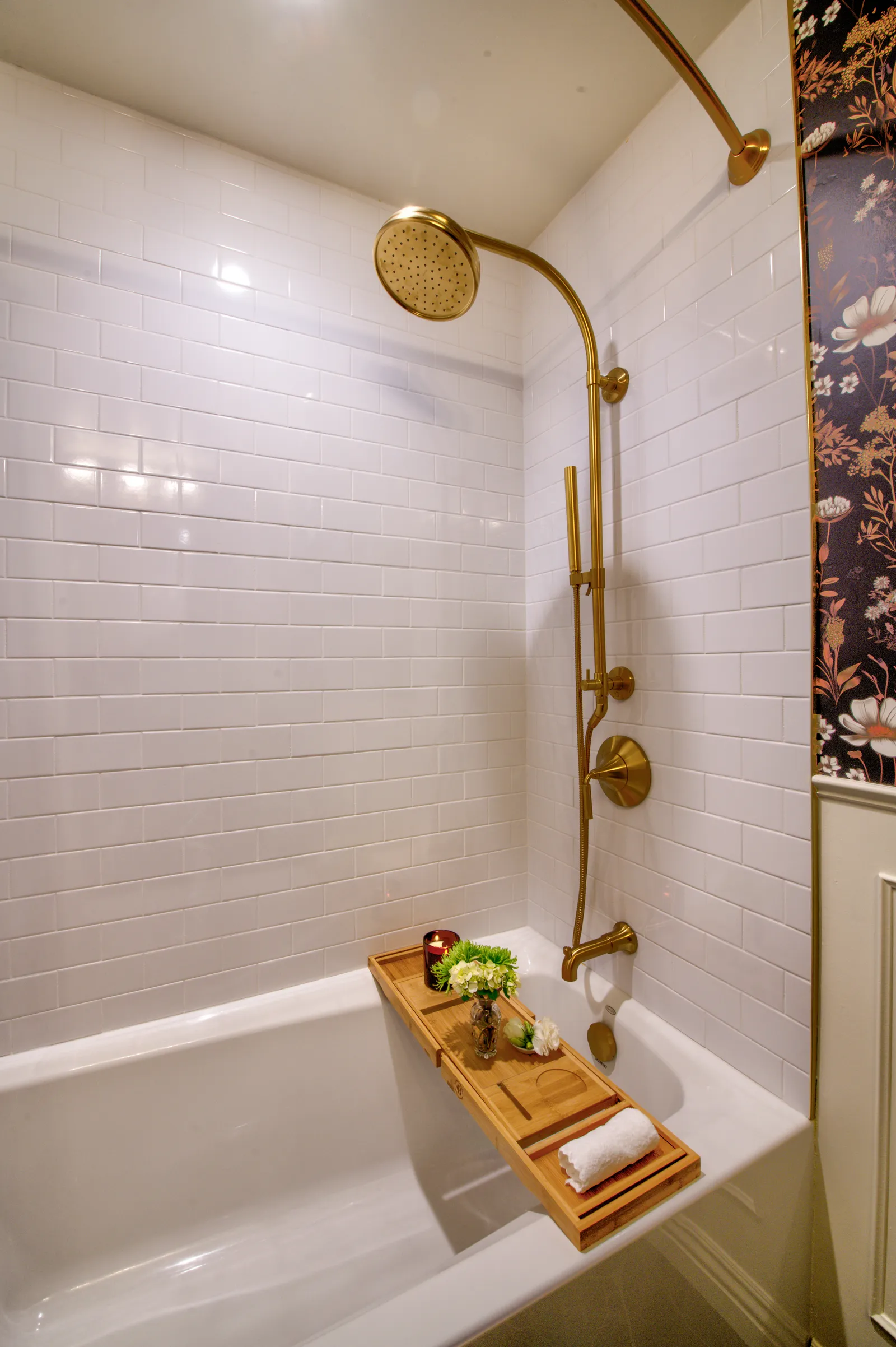Tub and shower with white subway tile, brass rain shower fixtures, and dark floral wallpaper.