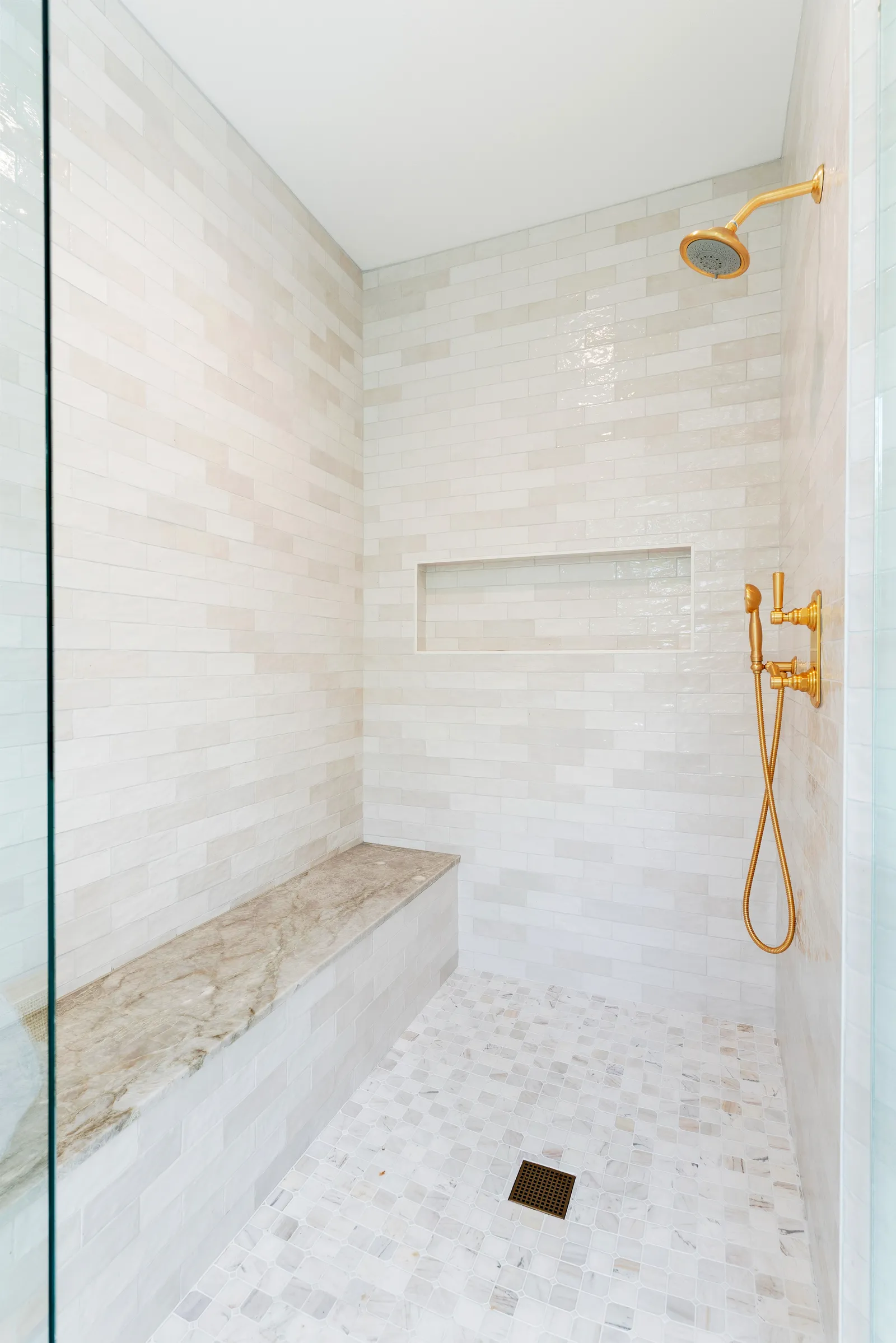 A walk-in shower with glossy cream subway tile, a marble-topped bench, mosaic stone floor, and brushed gold fixtures.