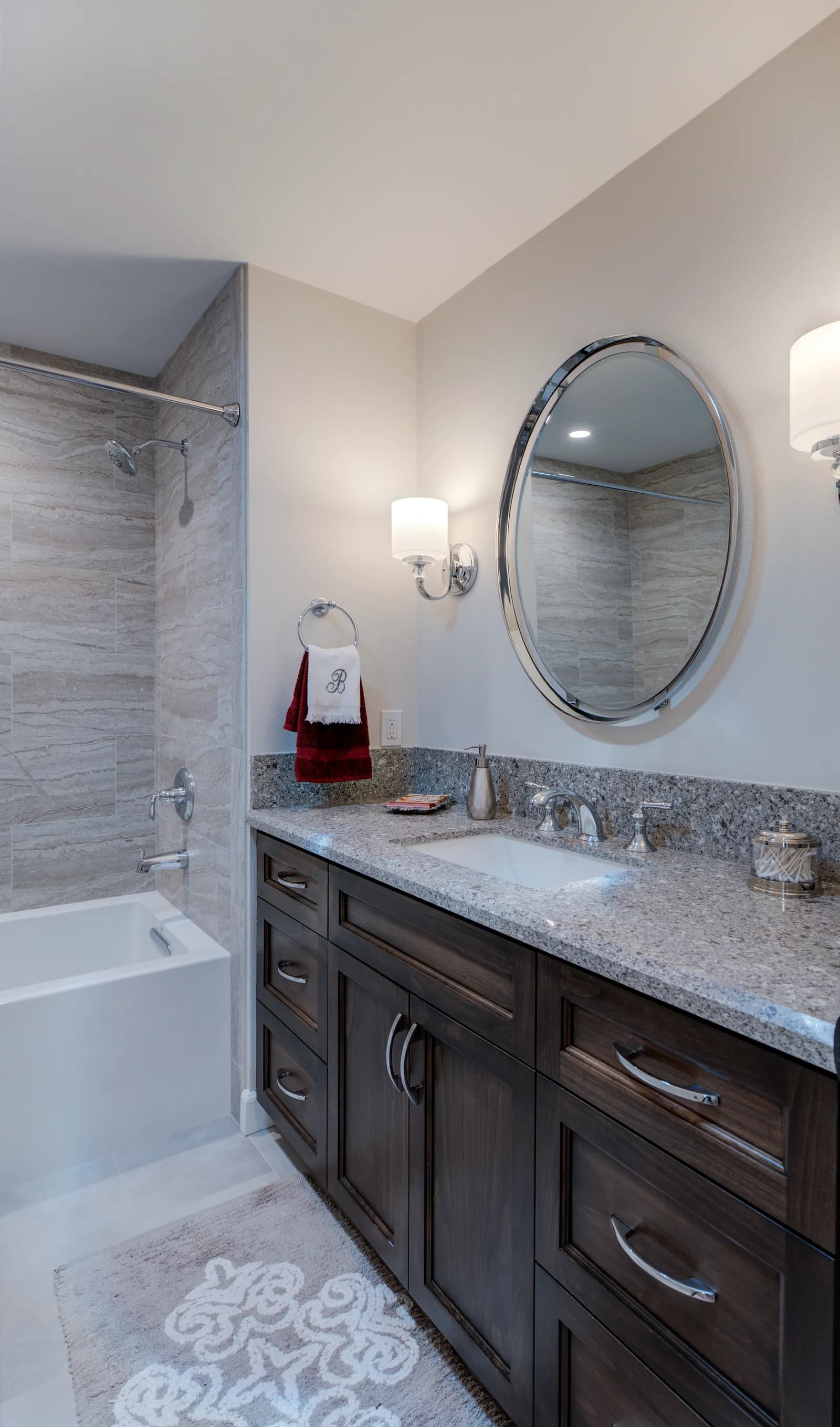 A bathroom with a dark wood vanity, speckled granite counter, chrome oval mirror, sconces, and a tub-shower with stone-look tile.