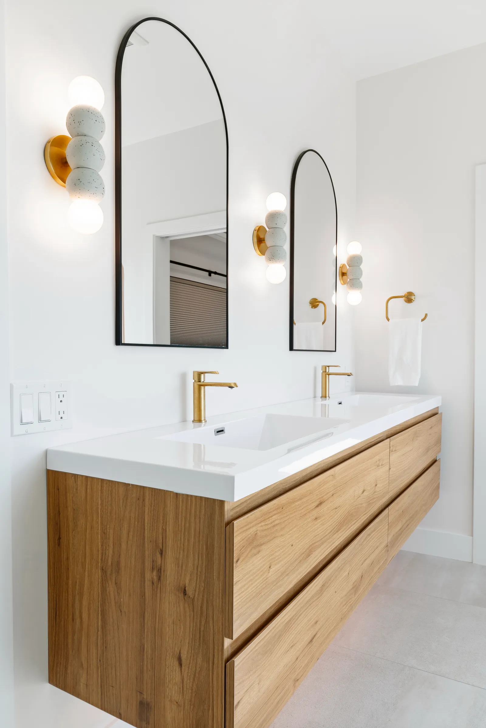 Floating white oak double vanity with arched mirrors, brass faucets, and sculptural globe sconces.