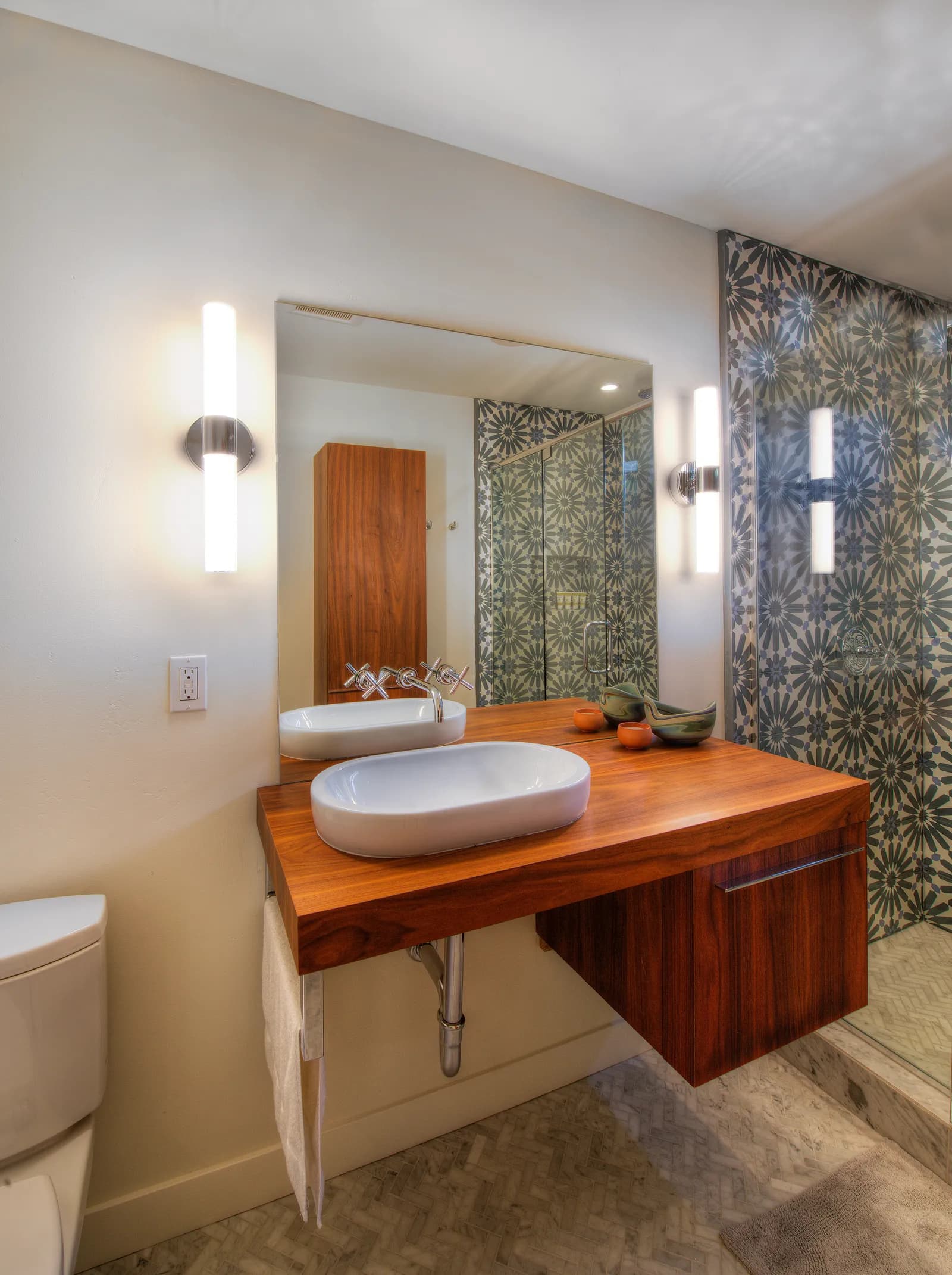 Bathroom with a floating walnut vanity, oval vessel sinks, wall-mounted cross-handle faucets, and a patterned tile shower wall.