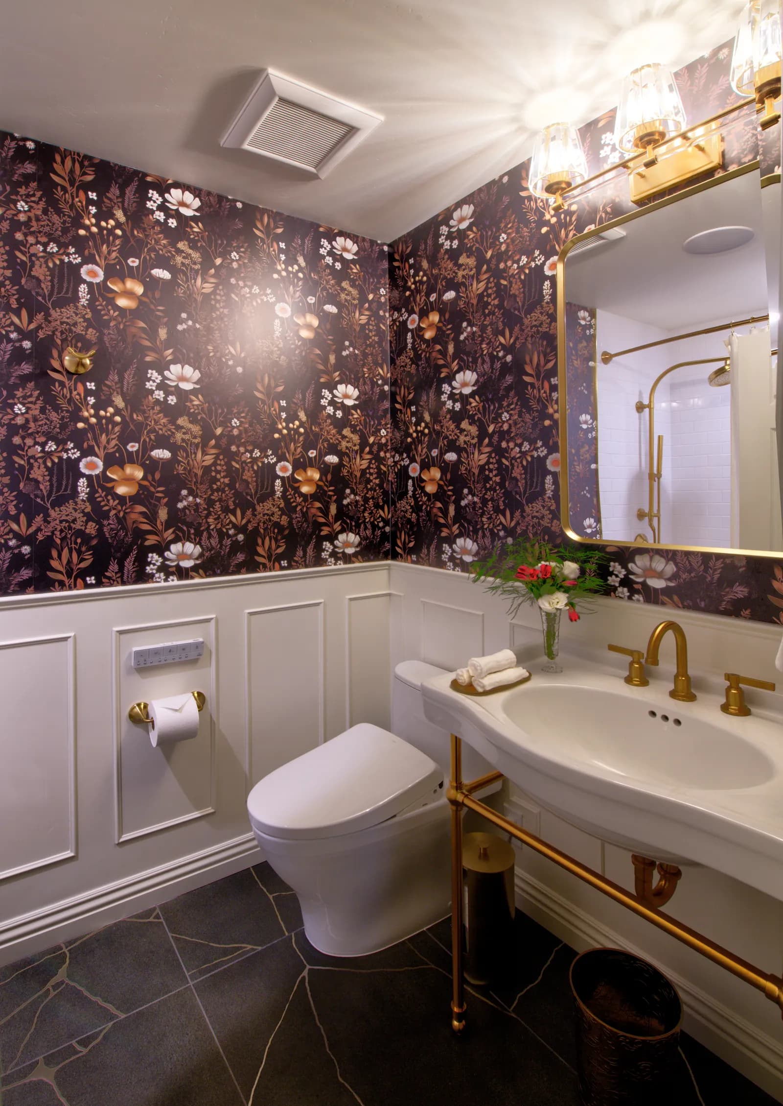 Elegant powder room with dark floral wallpaper, white wainscoting, brass console sink, and a brass-framed mirror.
