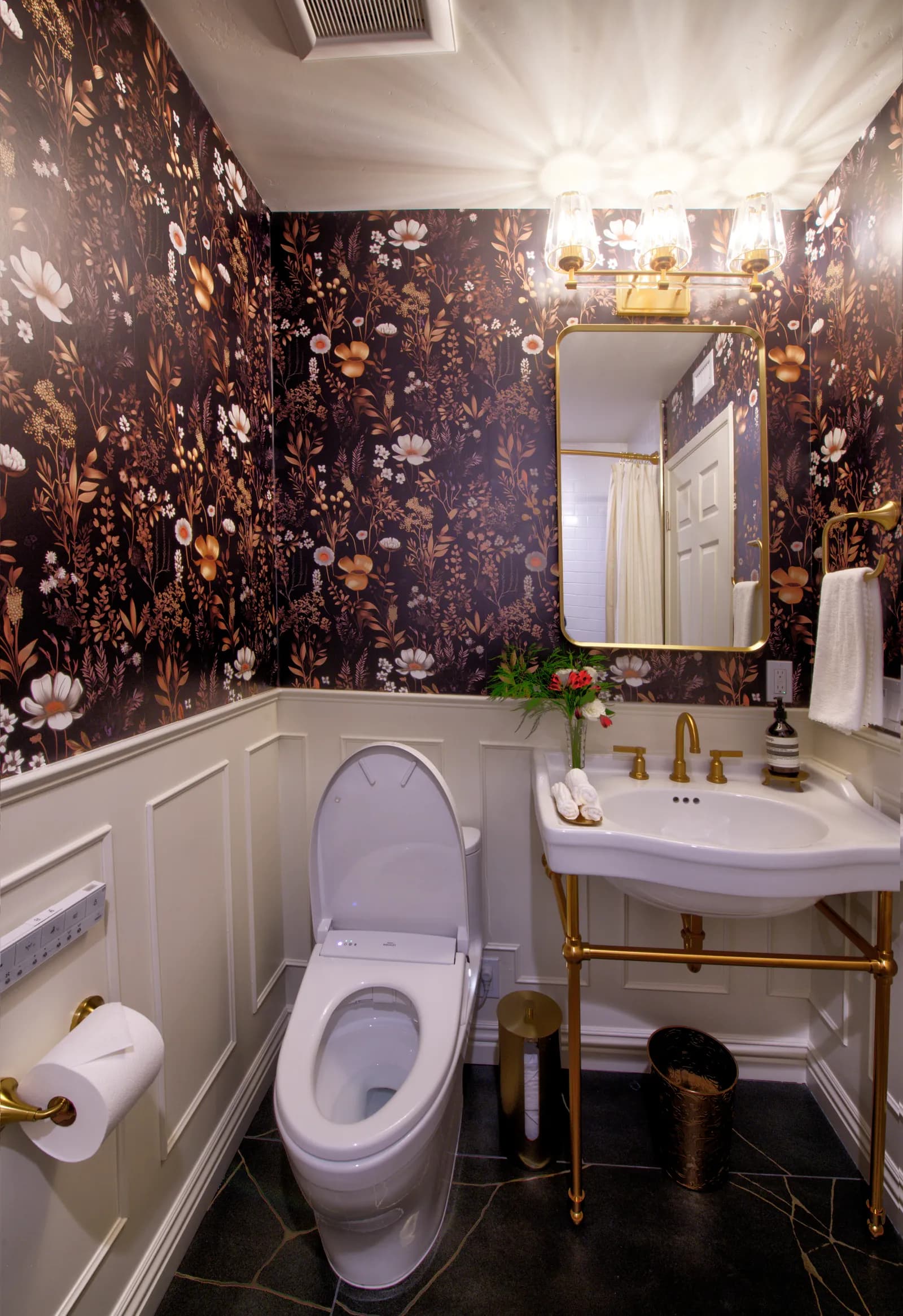 A powder room with dark floral wallpaper, cream wainscoting, a brass console sink, and brass light fixtures.