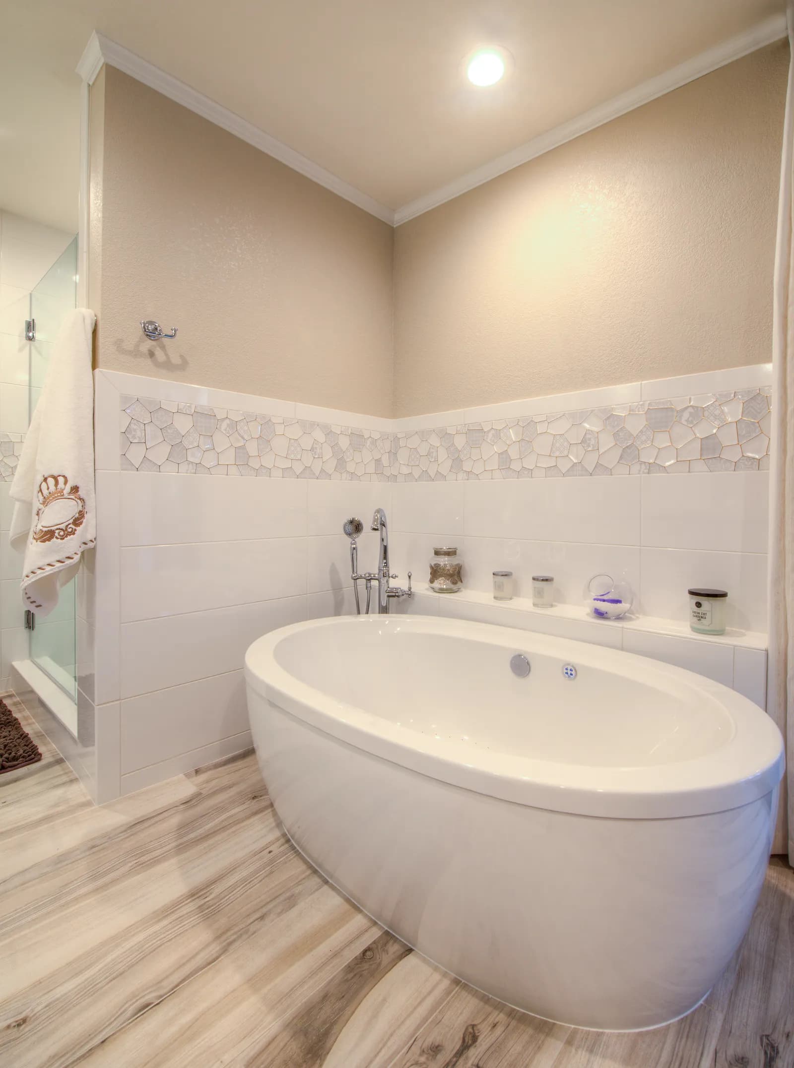 Bathroom with a freestanding oval soaking tub, wood-look tile floor, and a mosaic accent border above white wall tile.