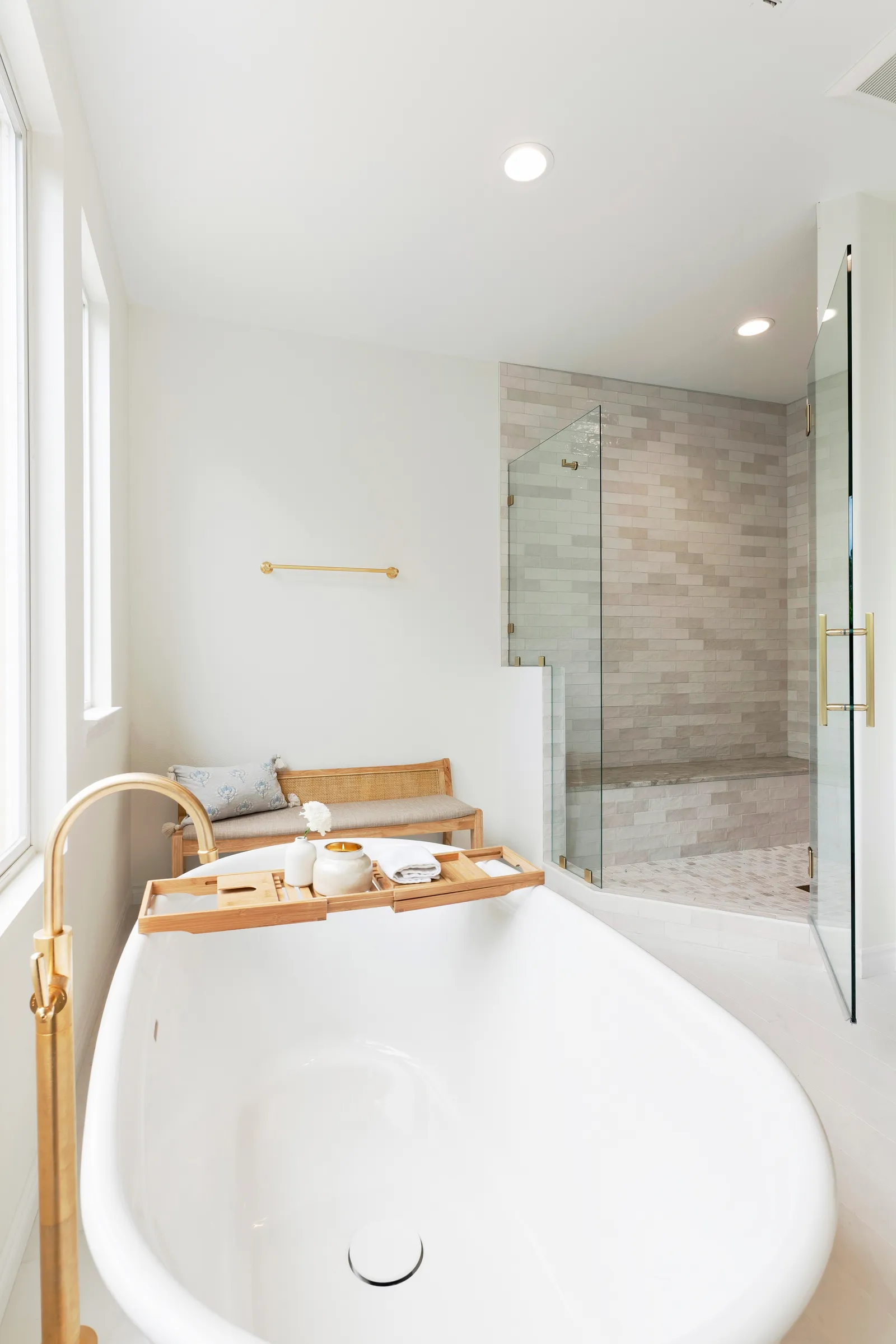 A bathroom with a white freestanding tub, brass floor-mounted filler, bamboo tray and glass-enclosed tile shower.