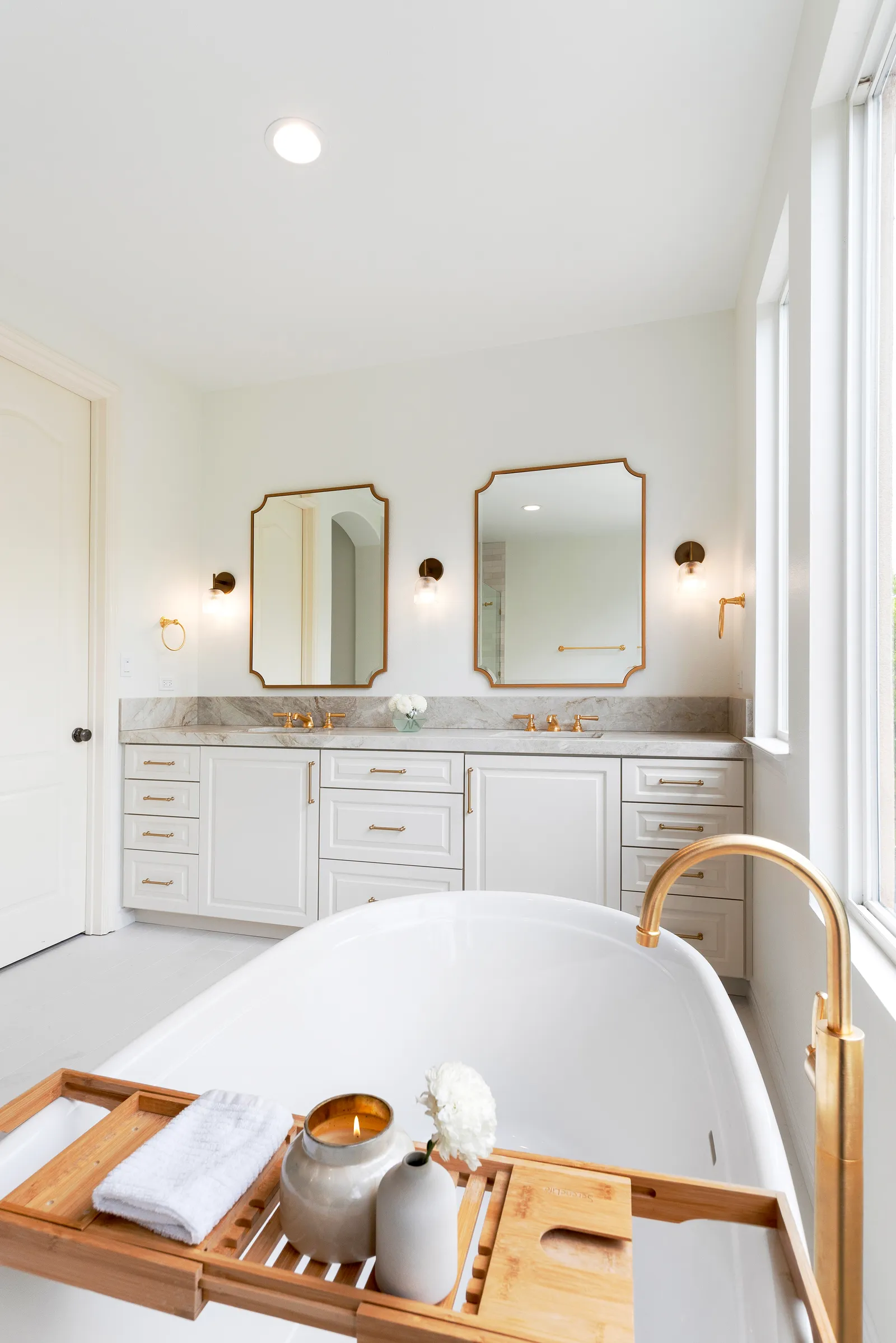 A bright bathroom with a freestanding white tub, brass floor-mounted filler, double vanity, and gold-framed mirrors.