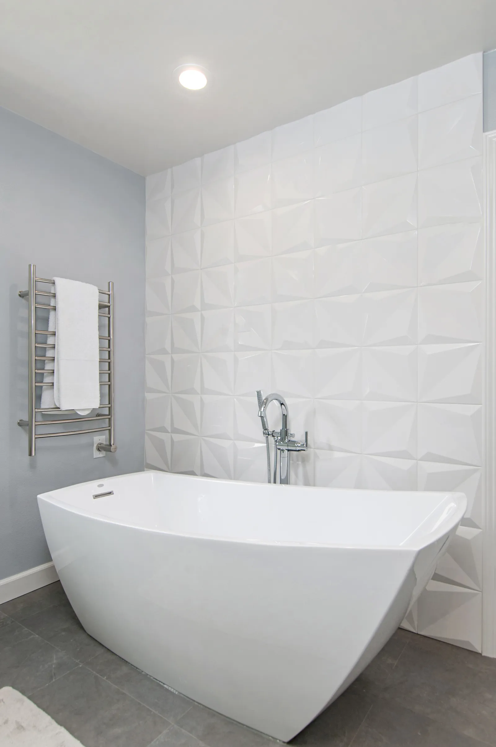 A freestanding white tub against a sculpted geometric tile wall with a floor-mounted chrome filler.