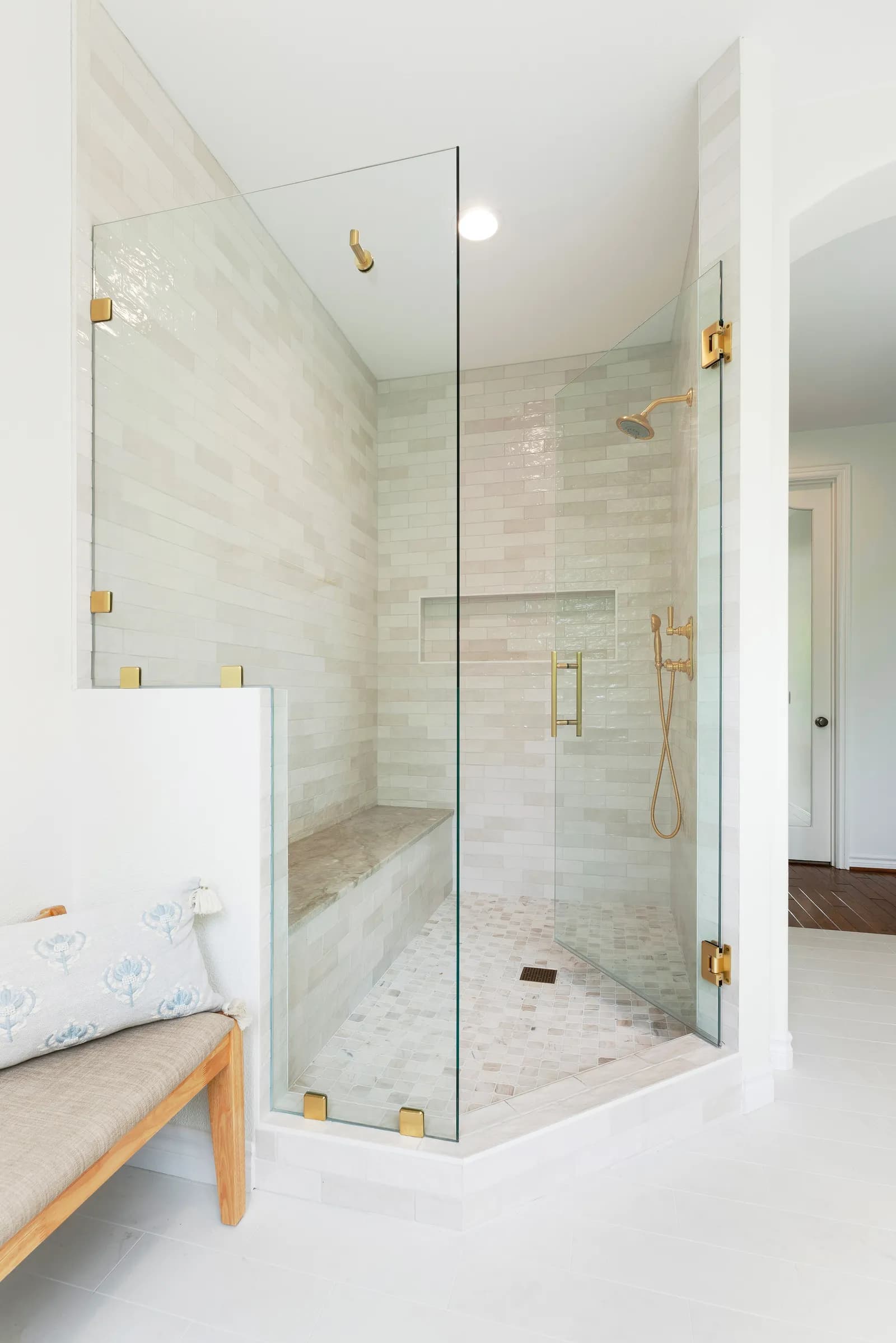 A frameless glass corner shower with glossy white tile, brass fixtures, a stone bench, and mosaic floor.