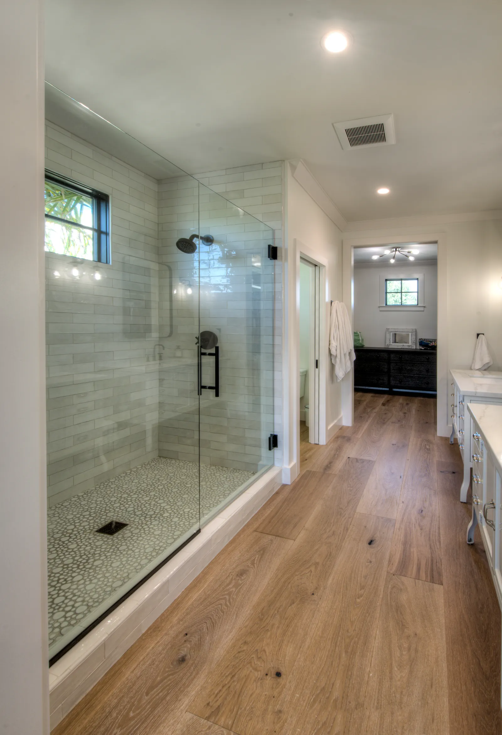 A long master bathroom with a frameless glass shower, pale green tile, pebble shower floor, and wide-plank oak flooring.