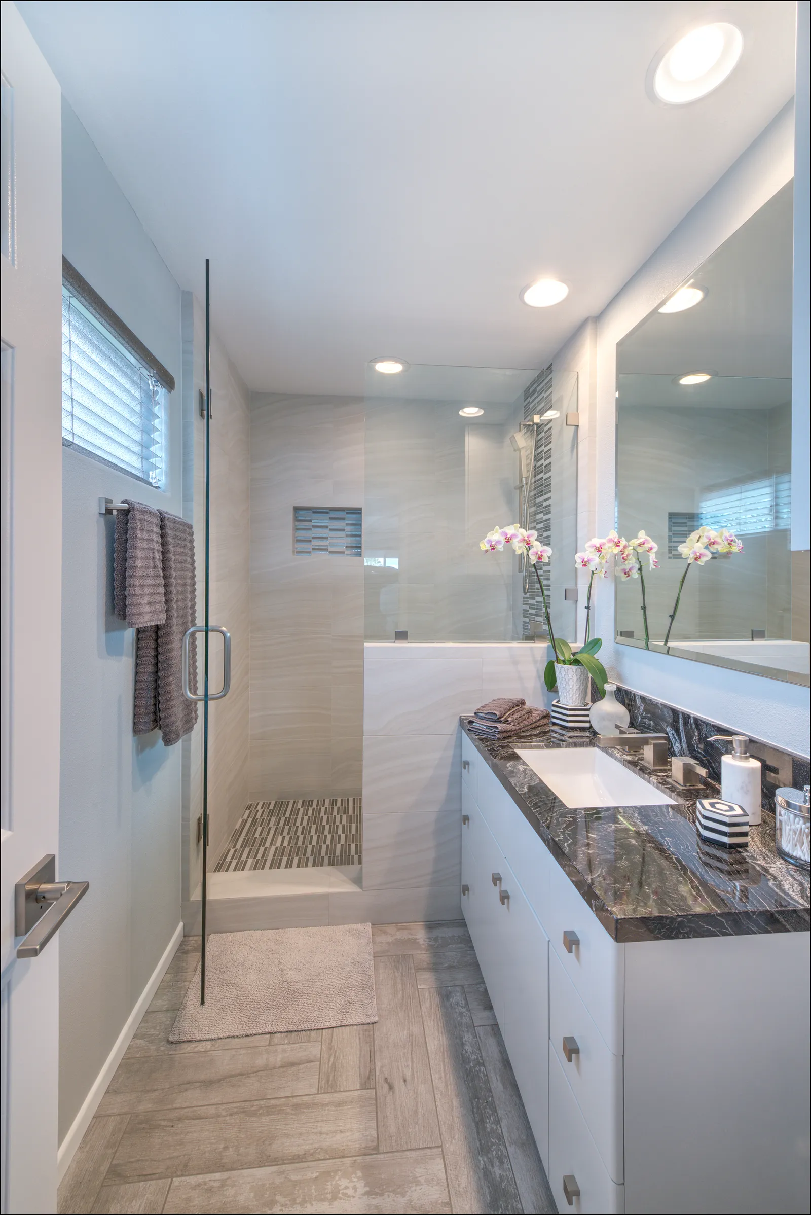 A modern bathroom with a glass walk-in shower, dark marble-topped vanity, and orchids by the mirror.