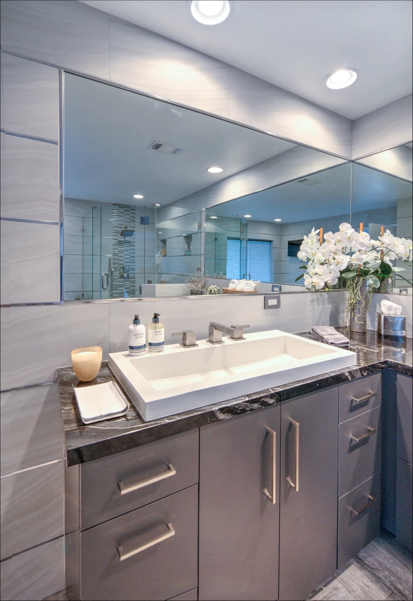 A modern bathroom vanity with gray flat-panel cabinets, a white trough sink, dark marbled countertop, and a wide frameless mirror.