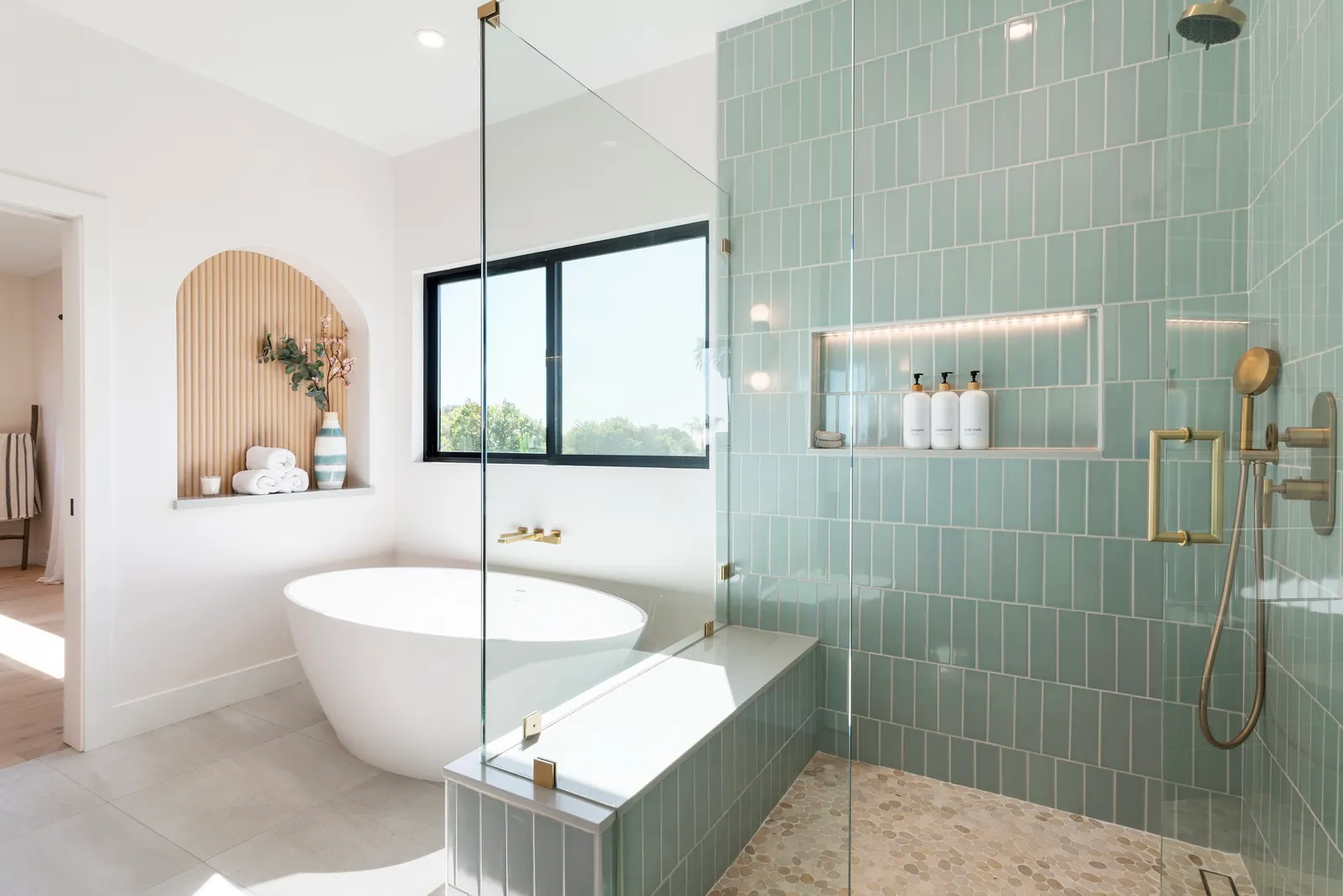 Bathroom with sage green vertical tile shower, glass enclosure, freestanding oval tub, and brass fixtures.