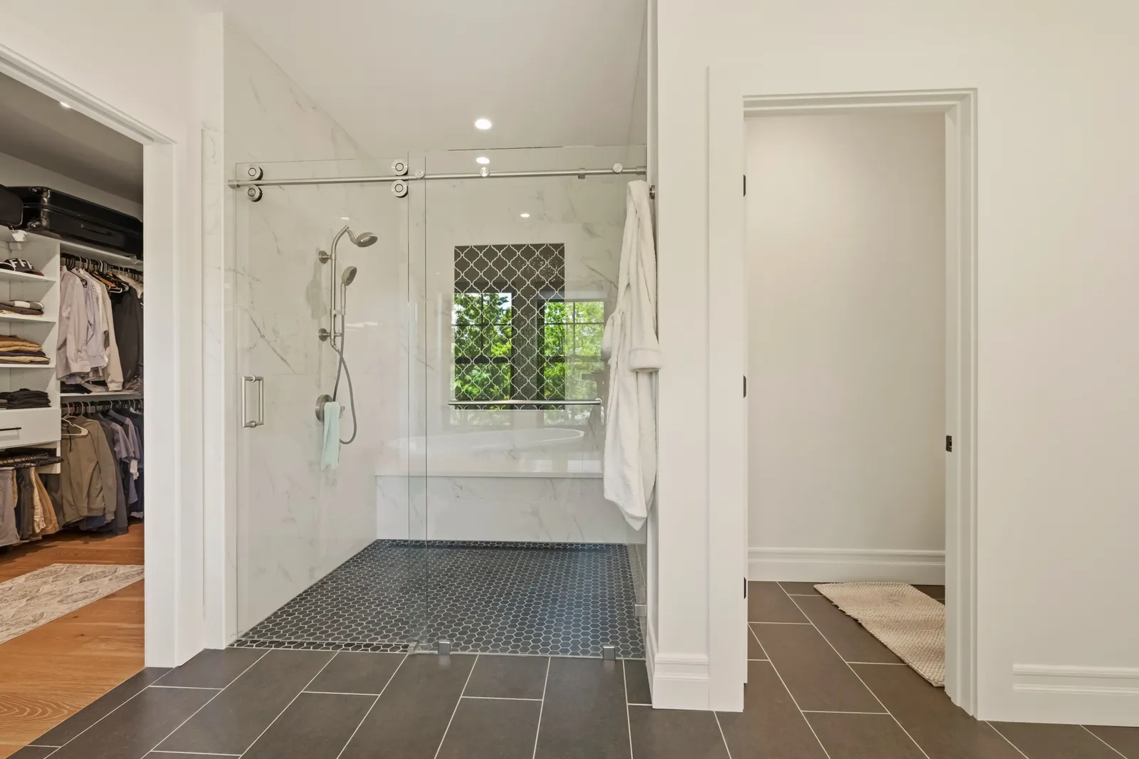 Marble-tiled walk-in shower with a sliding glass door, black hex tile floor, and a walk-in closet beyond.