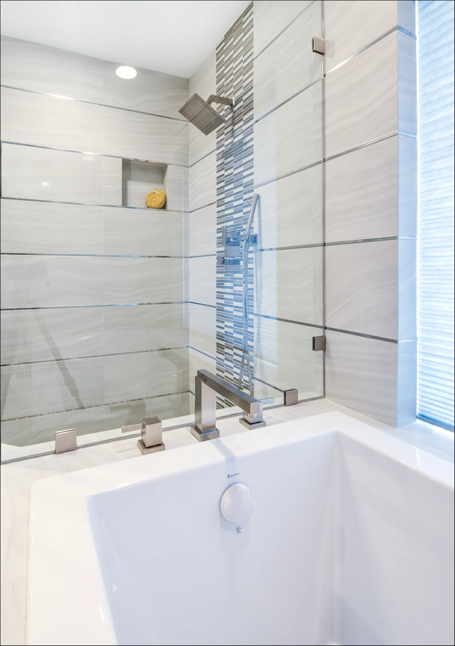 White rectangular soaking tub with brushed nickel deck-mount faucet beside a glass-enclosed shower with a mosaic accent stripe.
