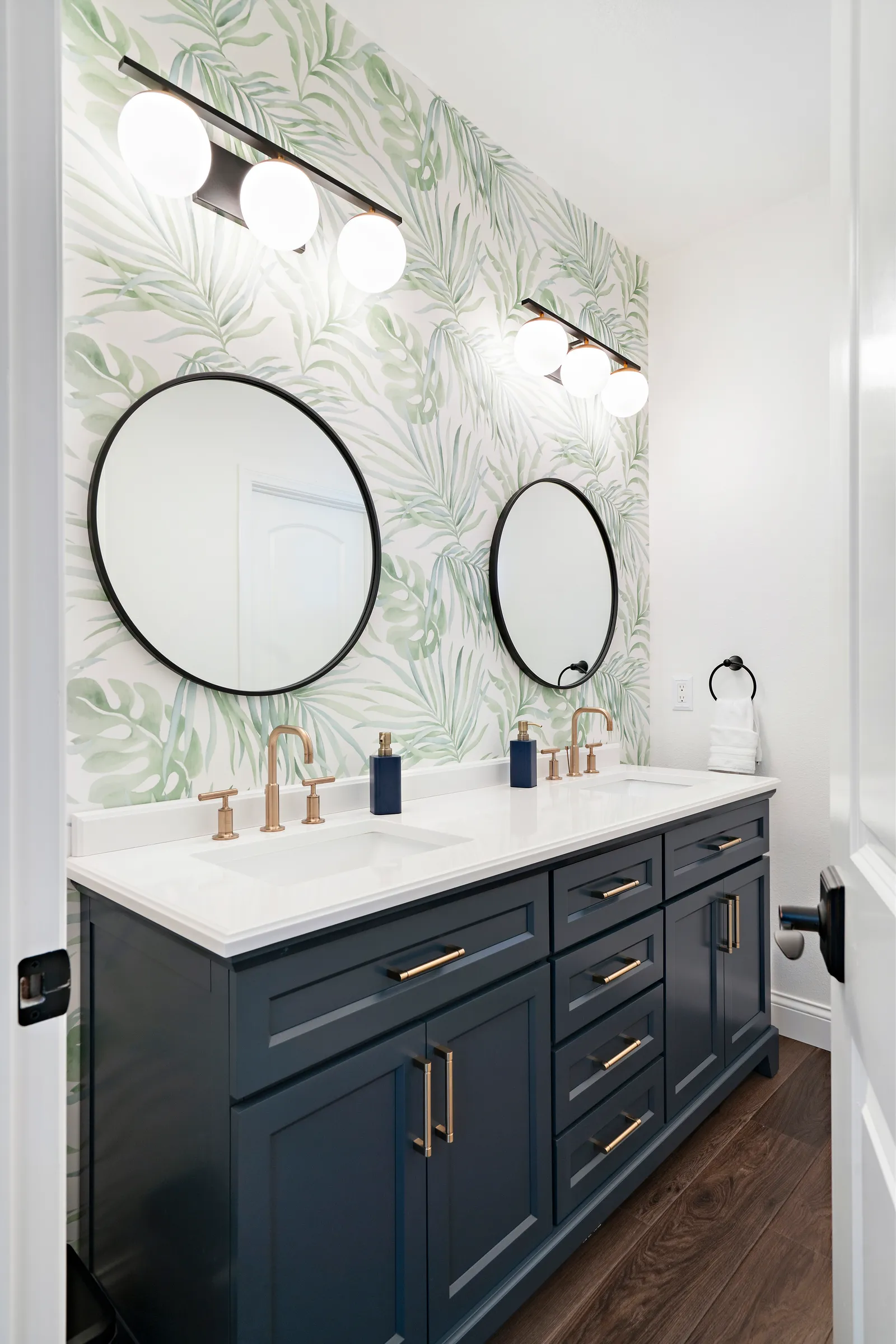 A bathroom with a navy double vanity, brass faucets, round black mirrors and green palm leaf wallpaper.