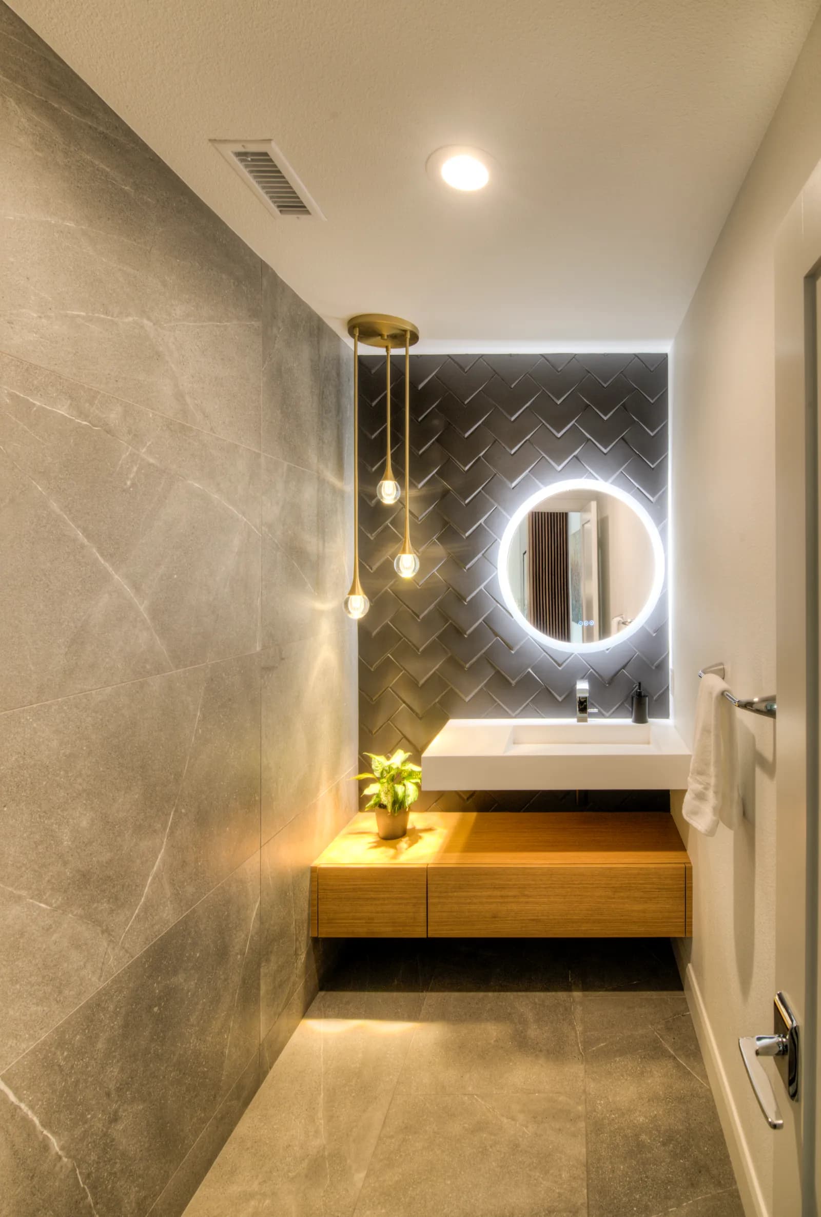 A modern powder room with a dark herringbone tile accent wall, backlit round mirror, floating wood vanity, and teardrop pendants.