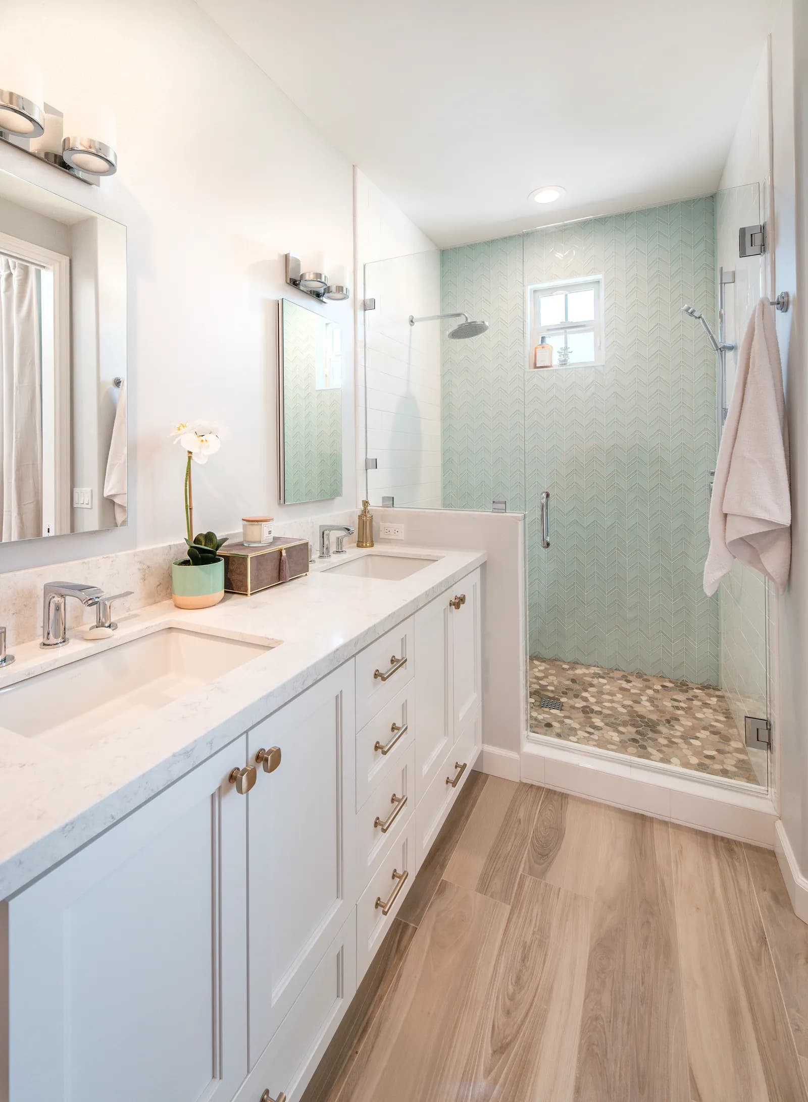Bathroom with white double vanity, brass hardware, and a glass shower with seafoam chevron tile and pebble floor.