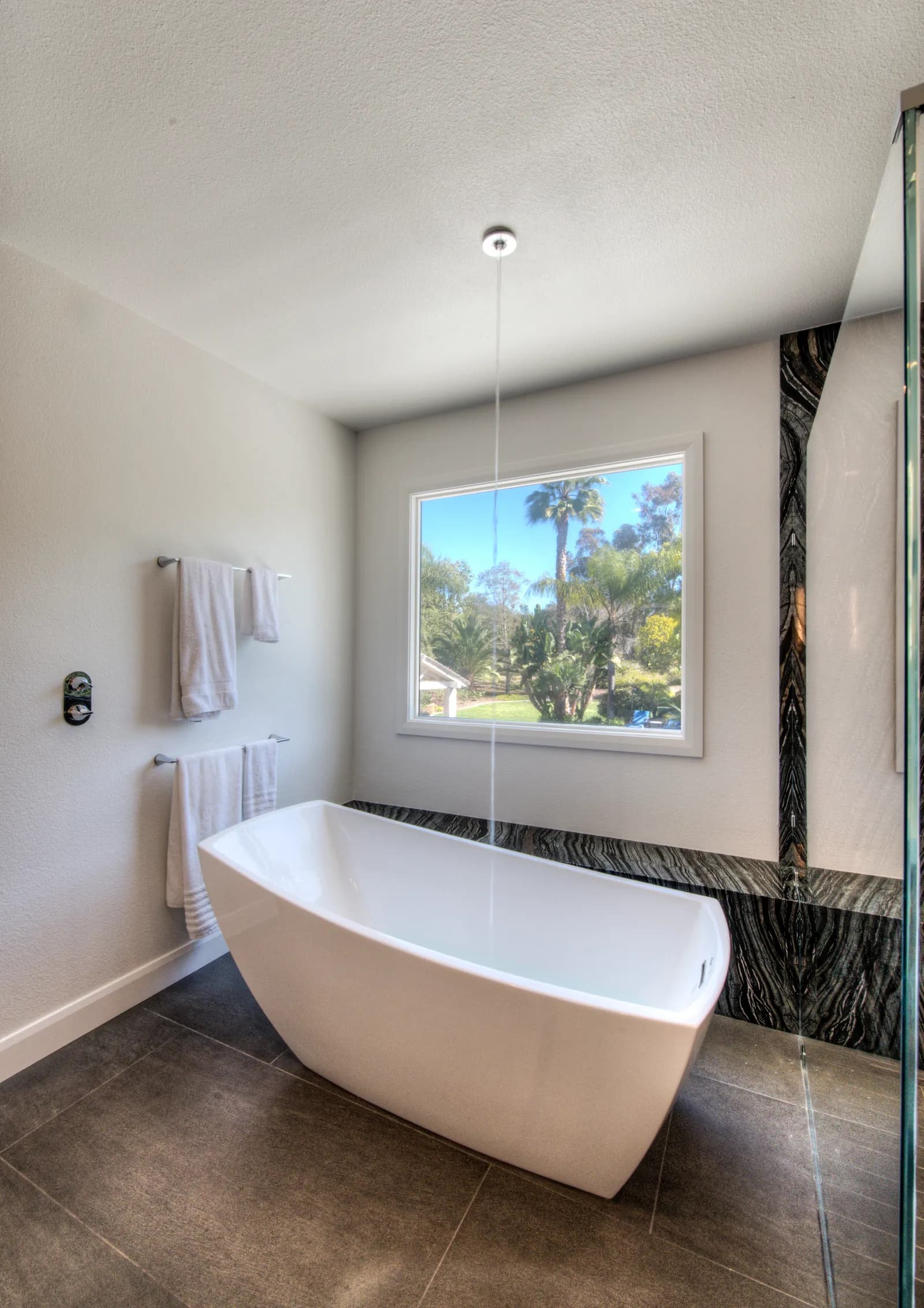 Modern white soaking tub filling from a ceiling-mounted tub filler beside a window with palm tree views and marble accents.