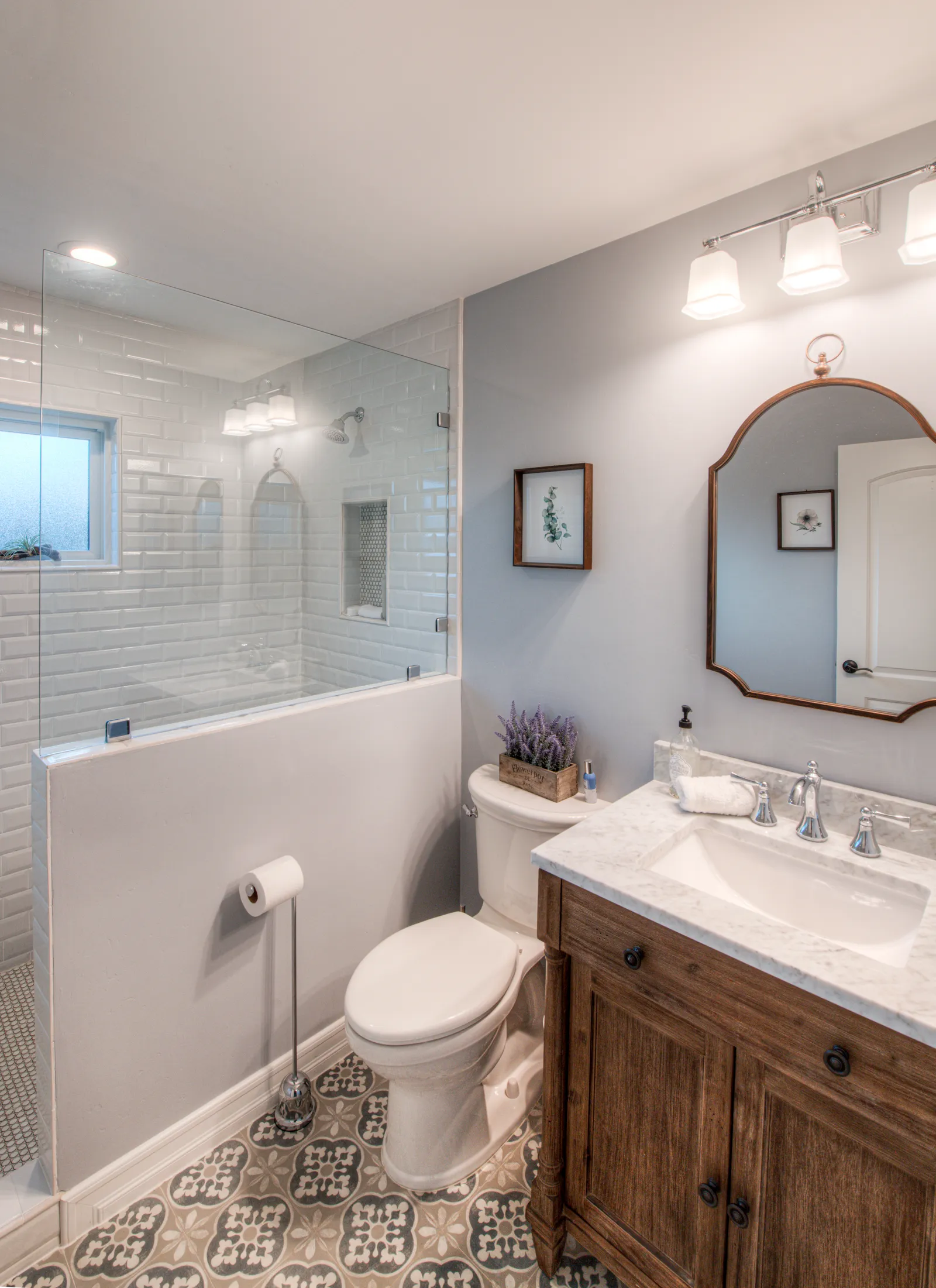 A bathroom with a beveled subway tile shower, arched copper-framed mirror, wood vanity, and patterned cement tile floor.