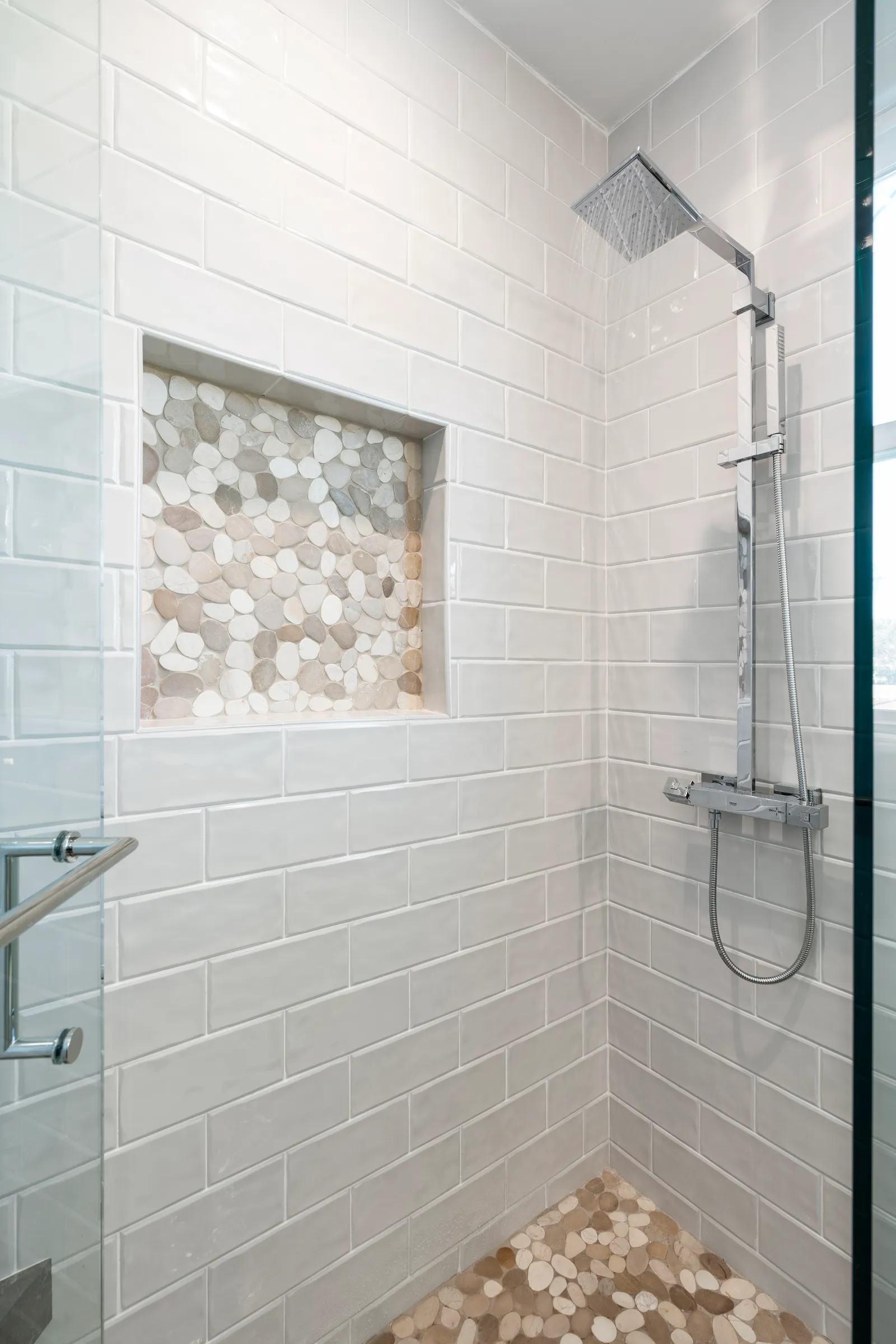 Walk-in shower with white subway tile, a pebble-filled niche, pebble floor, and a chrome rain shower column with water running.
