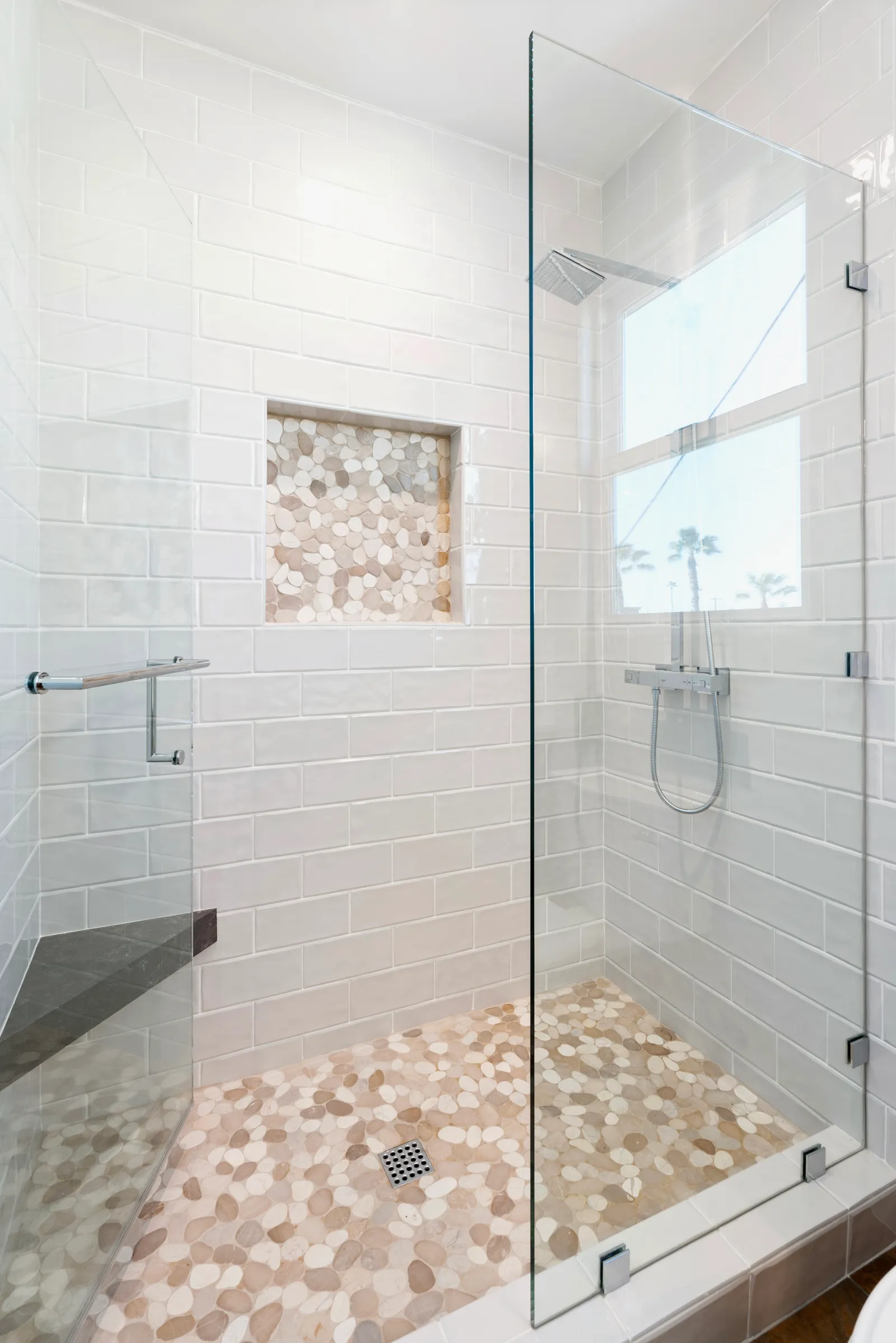 A walk-in shower with white subway tile, a pebble stone floor, frameless glass, and a bench.