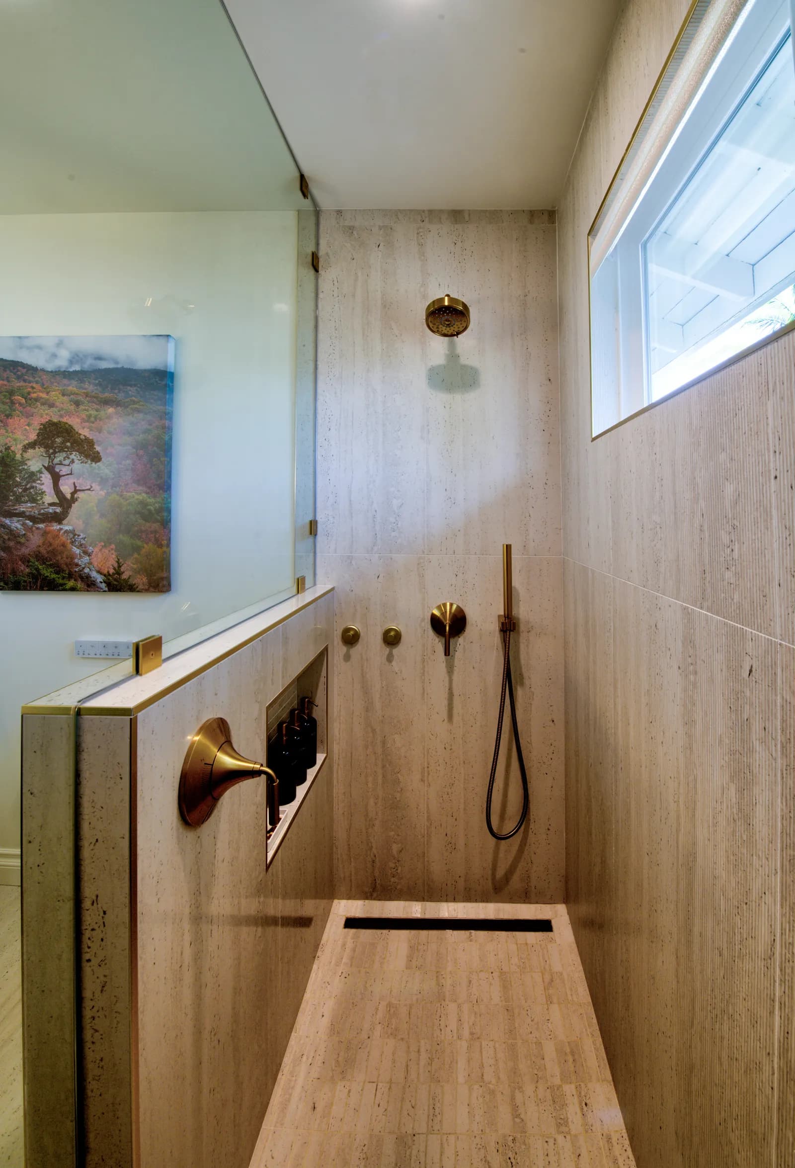 A walk-in shower with travertine-look tile walls, brass rain and handheld shower fixtures, and a linear drain.