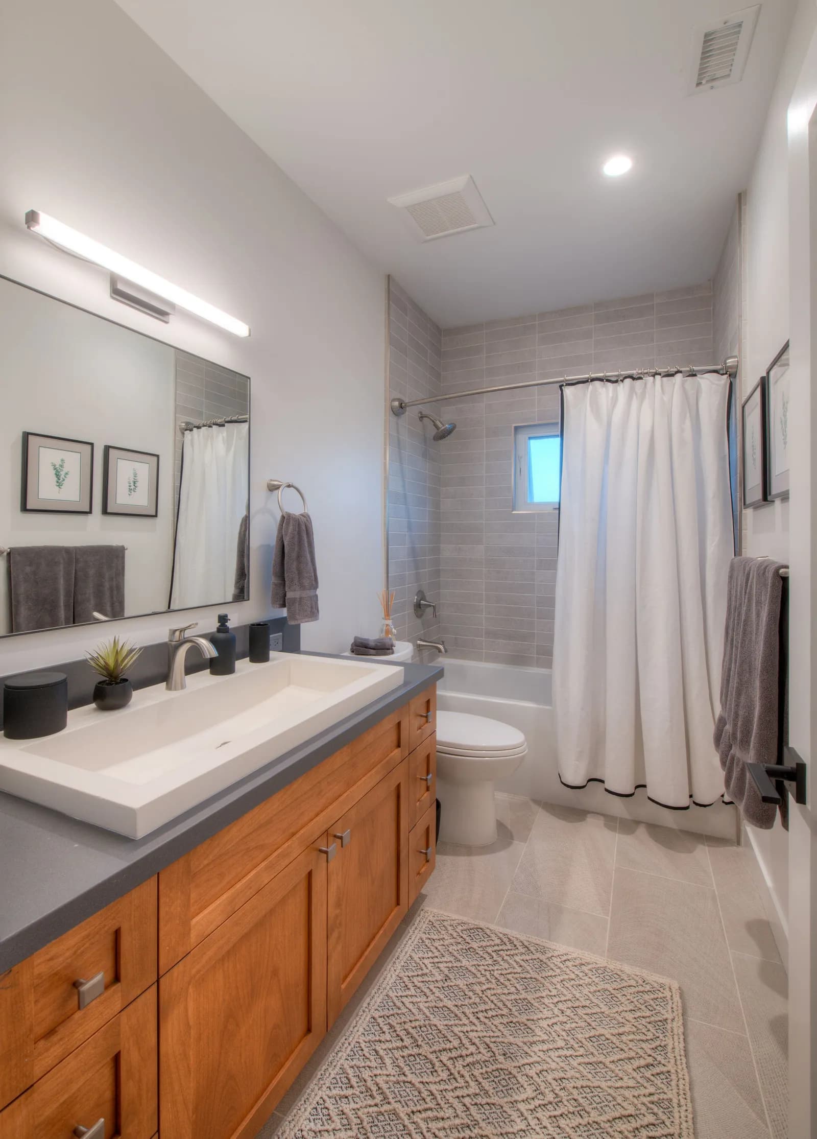Bathroom with a wide white trough sink on a cherry vanity, gray quartz counter, and a tiled tub-shower with white curtain.