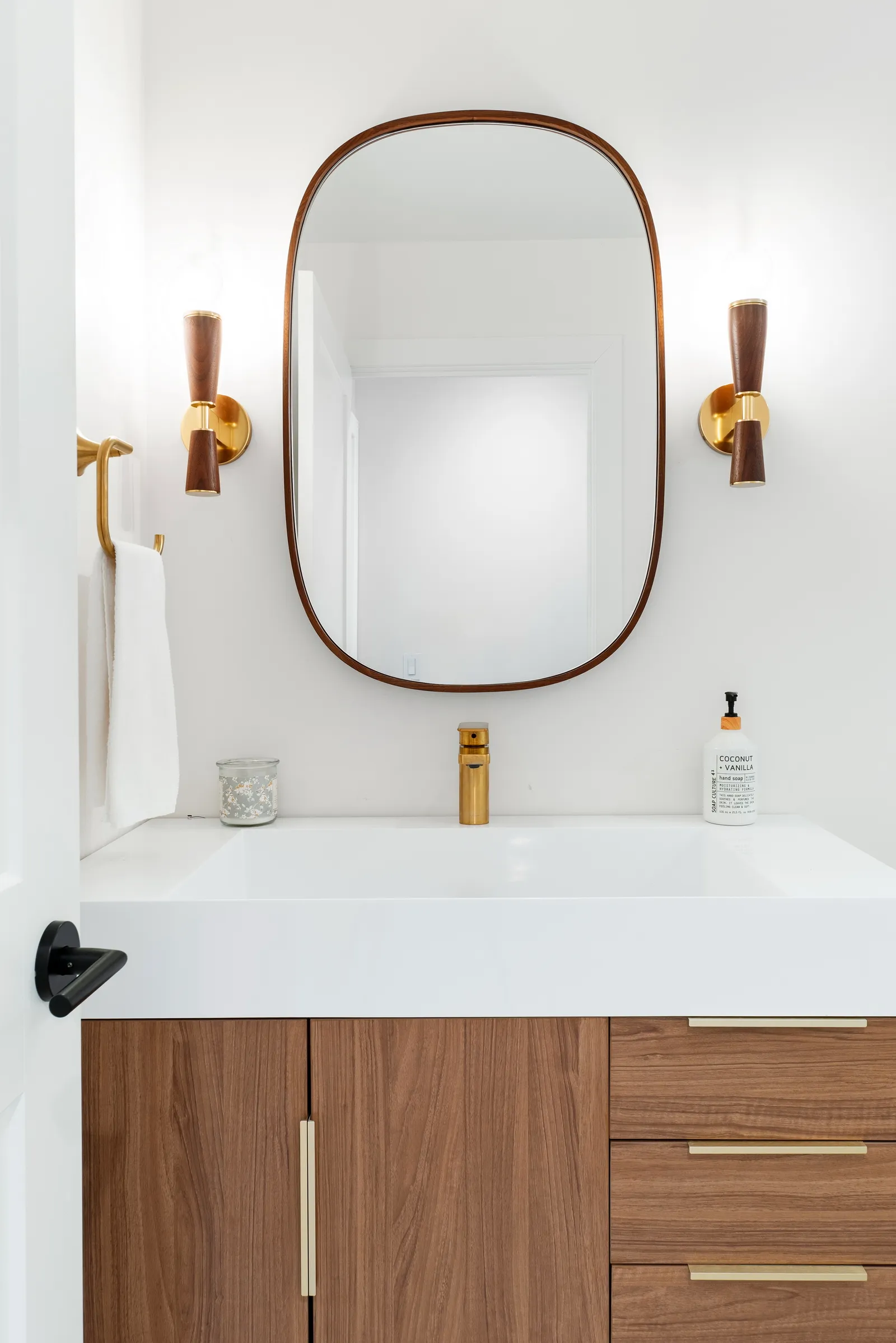 Bathroom vanity with walnut cabinets, white countertop sink, brass faucet, and wood-framed oval mirror.