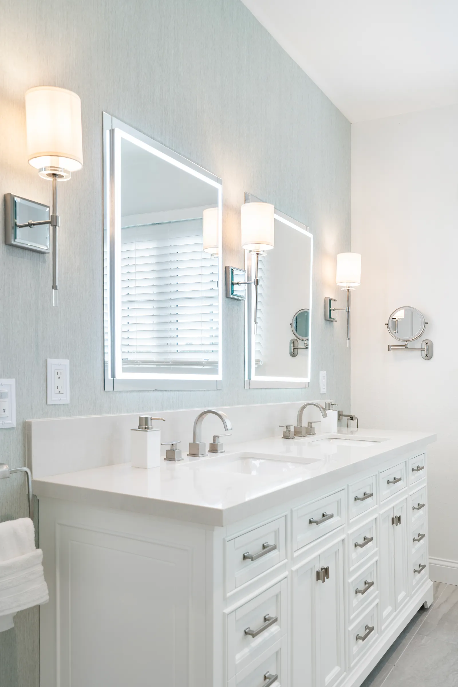 A bathroom with a white double vanity, brushed nickel faucets, LED-lit mirrors, and wall sconces.