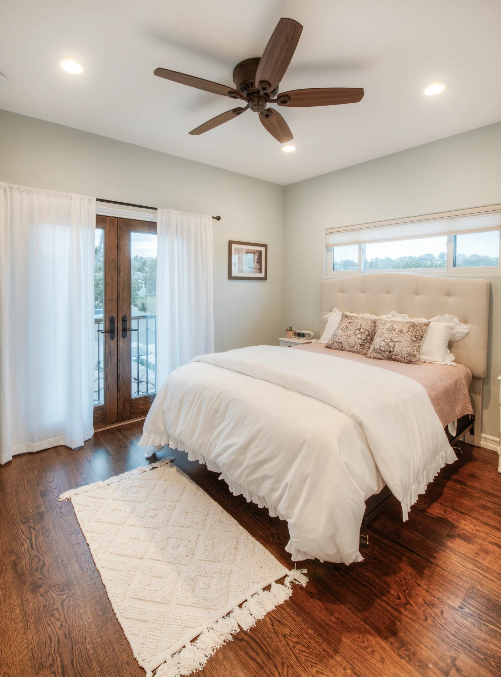 Bedroom with a tufted headboard, wood French doors to a balcony, ceiling fan, and hardwood floors.