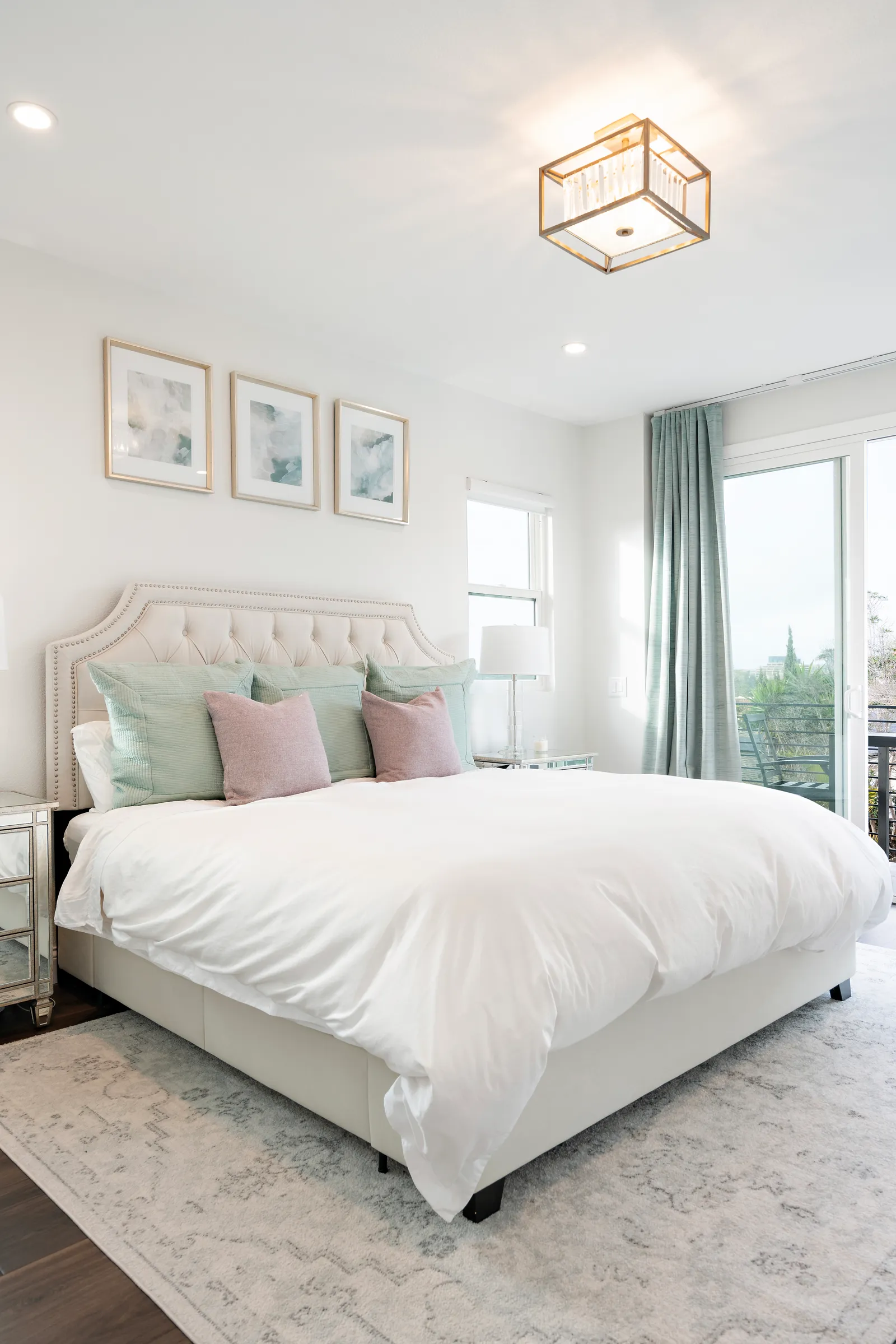 A bright bedroom with a cream tufted headboard, pastel pillows, seafoam curtains, and a sliding door to a balcony.