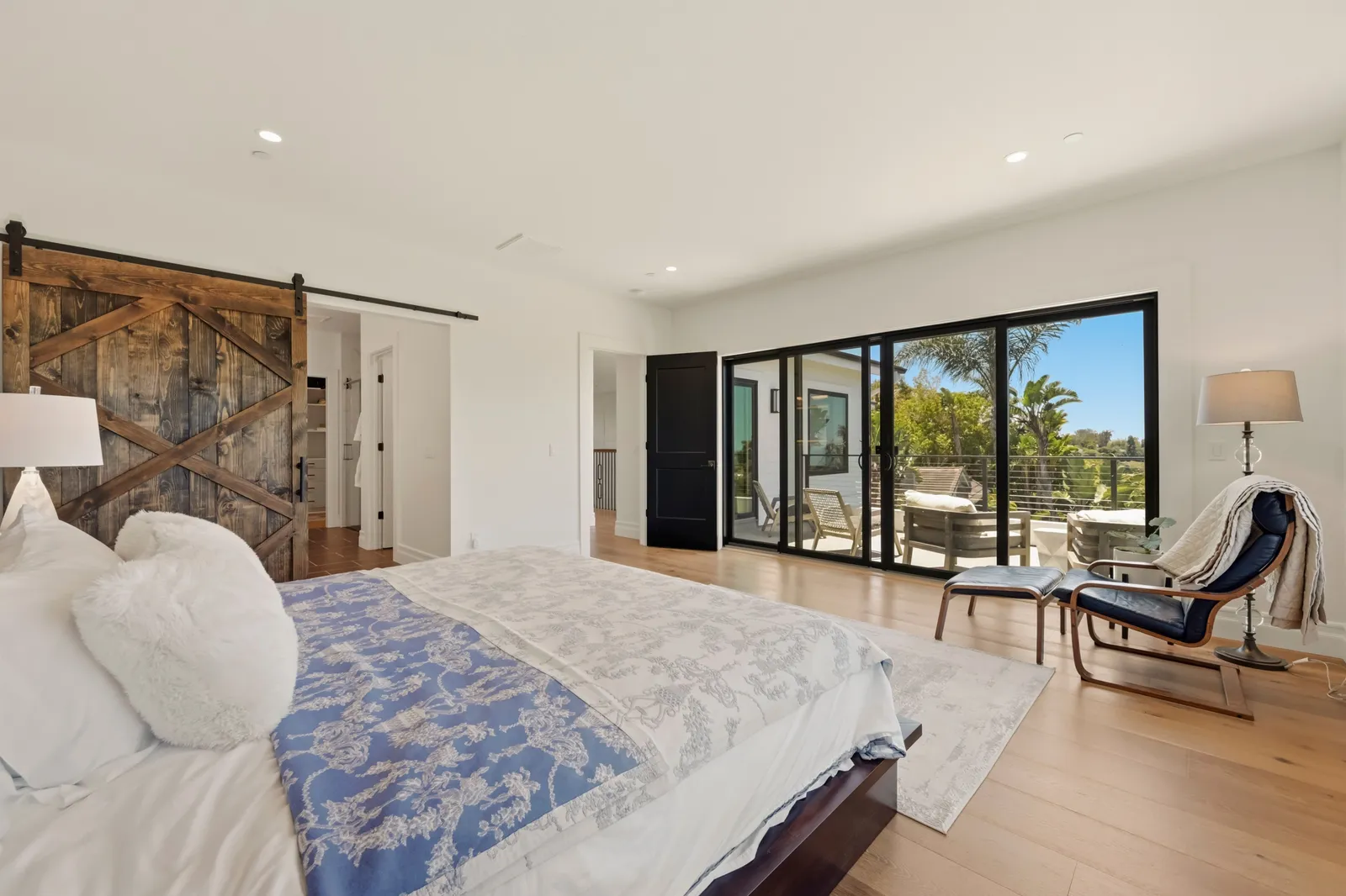 A primary bedroom with a rustic sliding barn door, blue and white bedding, and black-framed sliding doors opening to a deck with lounge seating.