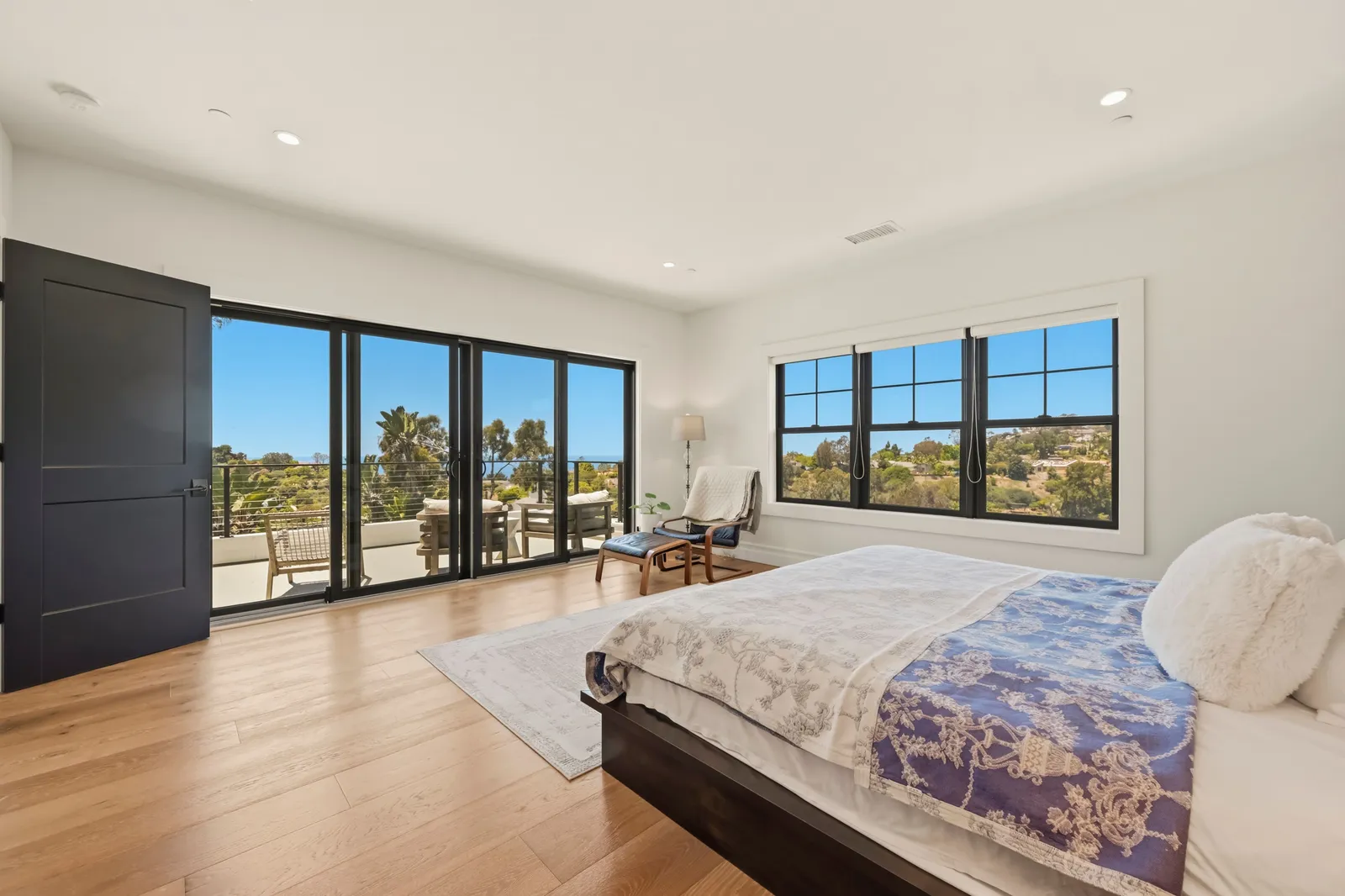 A spacious bedroom with wide sliding doors to a balcony, large windows, and a bed with a blue patterned coverlet.