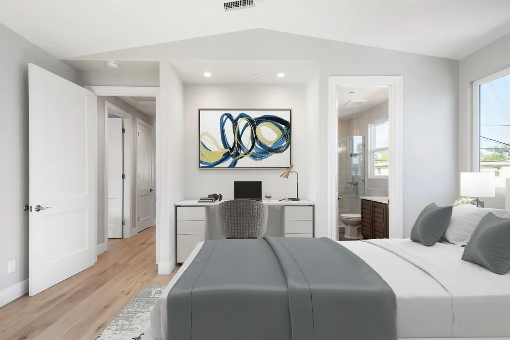 A staged bedroom with gray bedding, a built-in desk nook with abstract art, and a view into an ensuite bathroom.