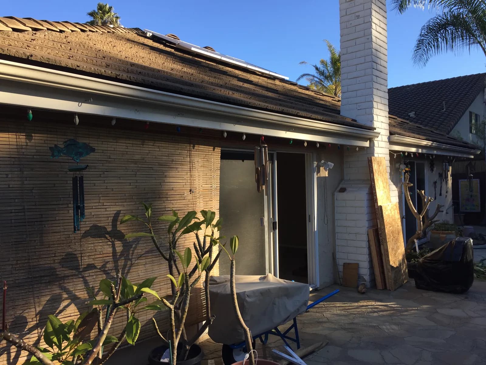The rear of a brick and stucco home with plywood sheets leaning against a white chimney and a covered wheelbarrow on the patio.