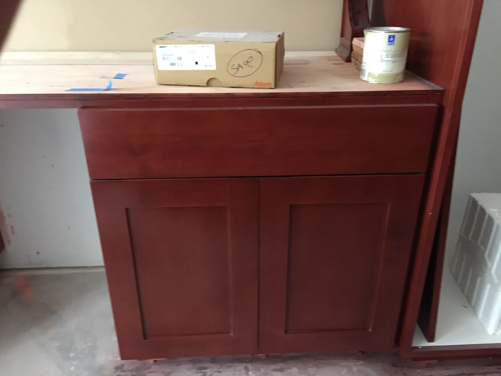 A cherry base cabinet staged during installation with a hardware box and primer can on the unfinished plywood top.