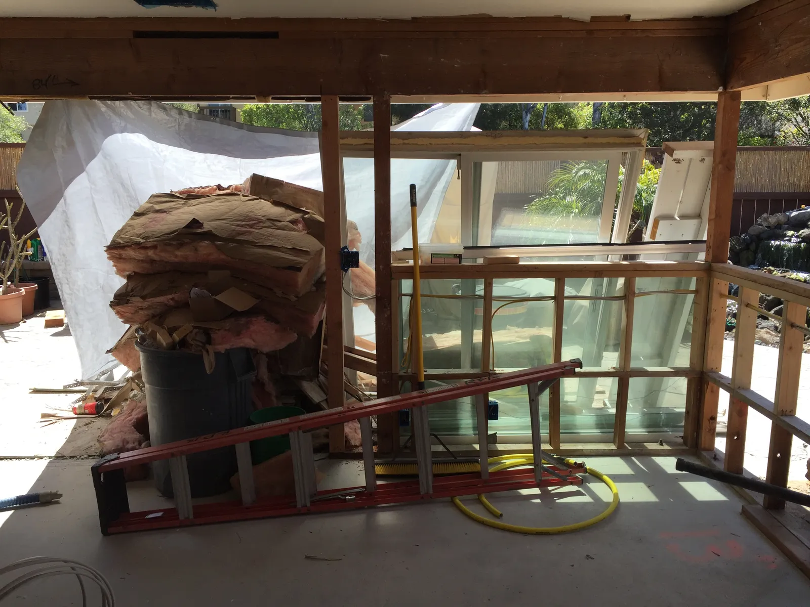 A remodel in progress with exposed wood framing, piled insulation, a ladder, tarps, and stacked window units.