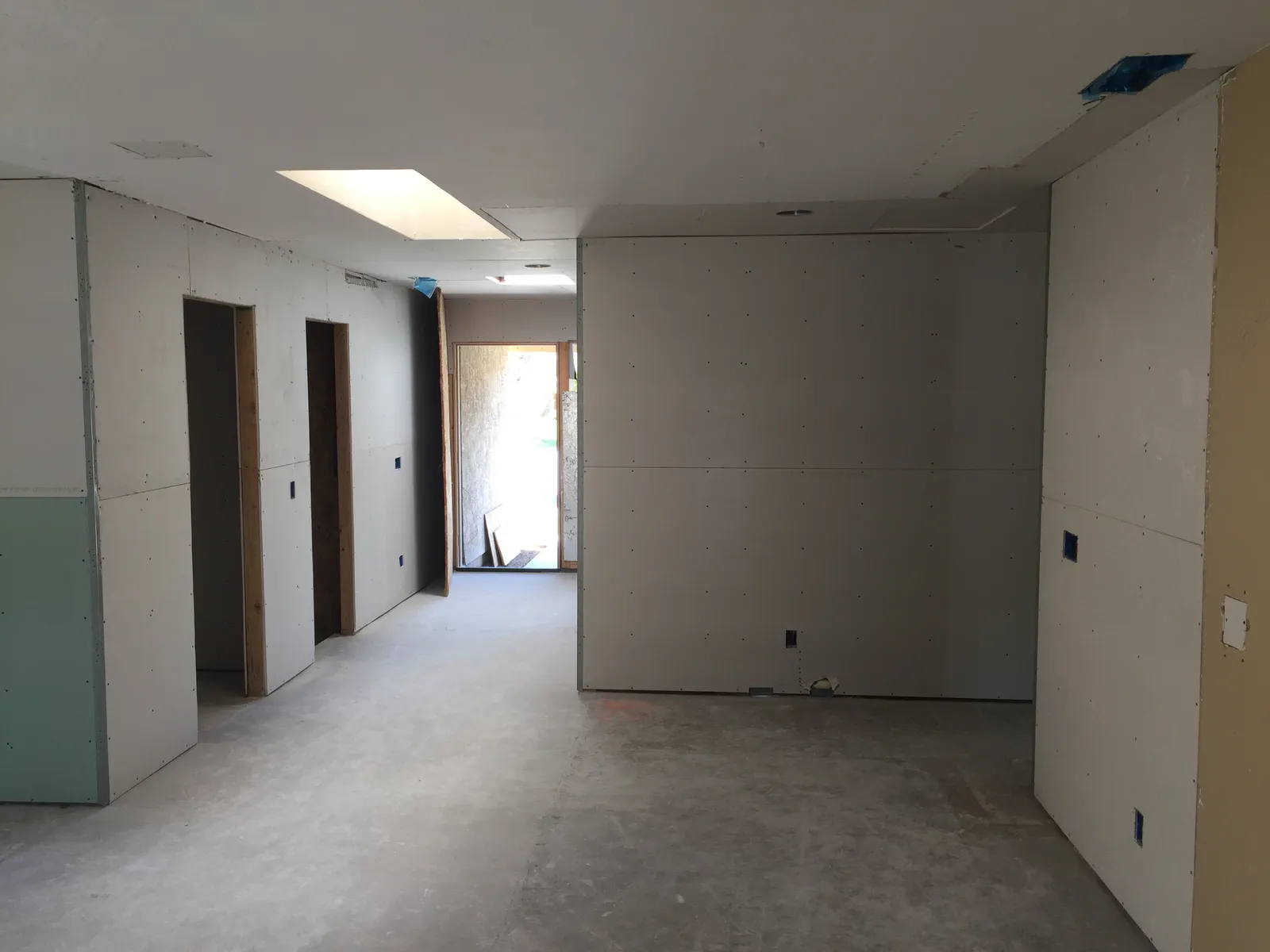 Interior remodel in progress with new drywall hung on walls and ceiling and sunlight coming through a skylight and open front door.