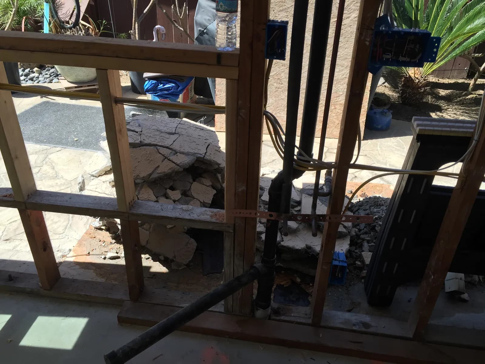 An open framed exterior wall during demolition showing exposed plumbing, wiring, and broken stucco.