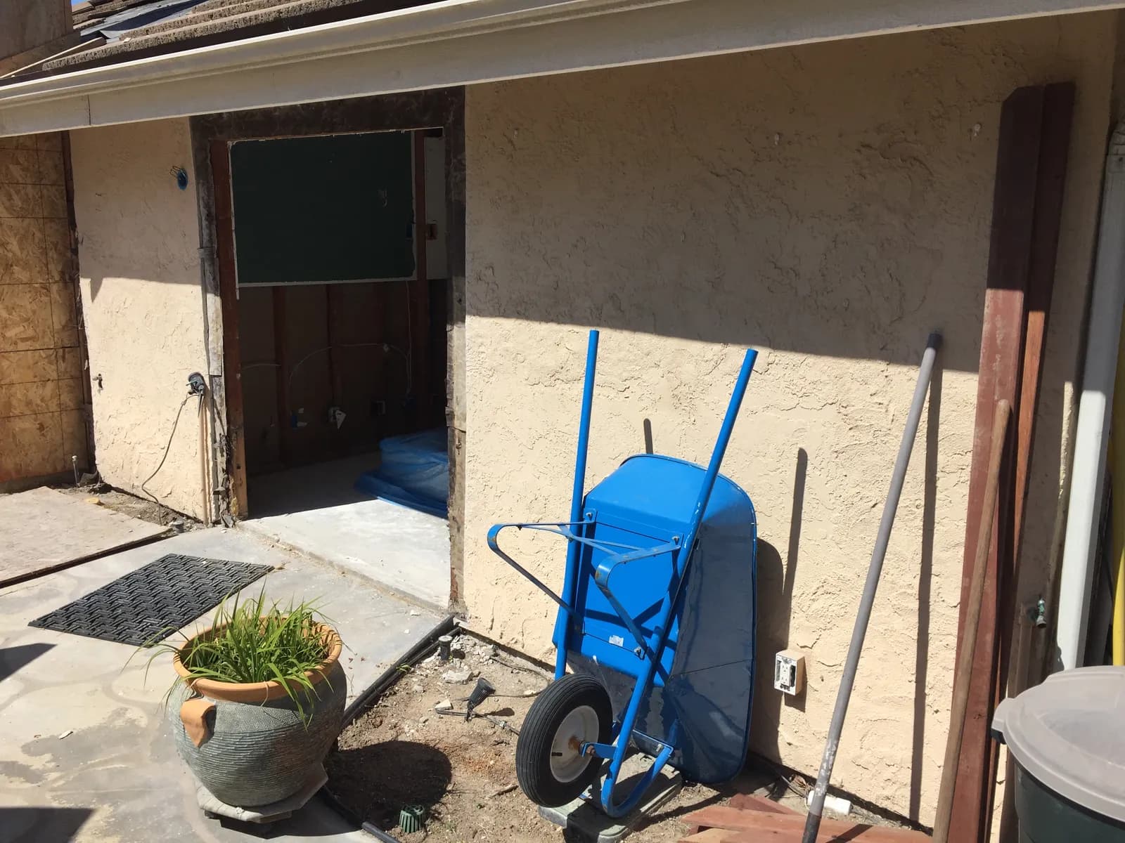 Stucco exterior during demolition with an opened wall cavity, a wheelbarrow, and tools leaning nearby.