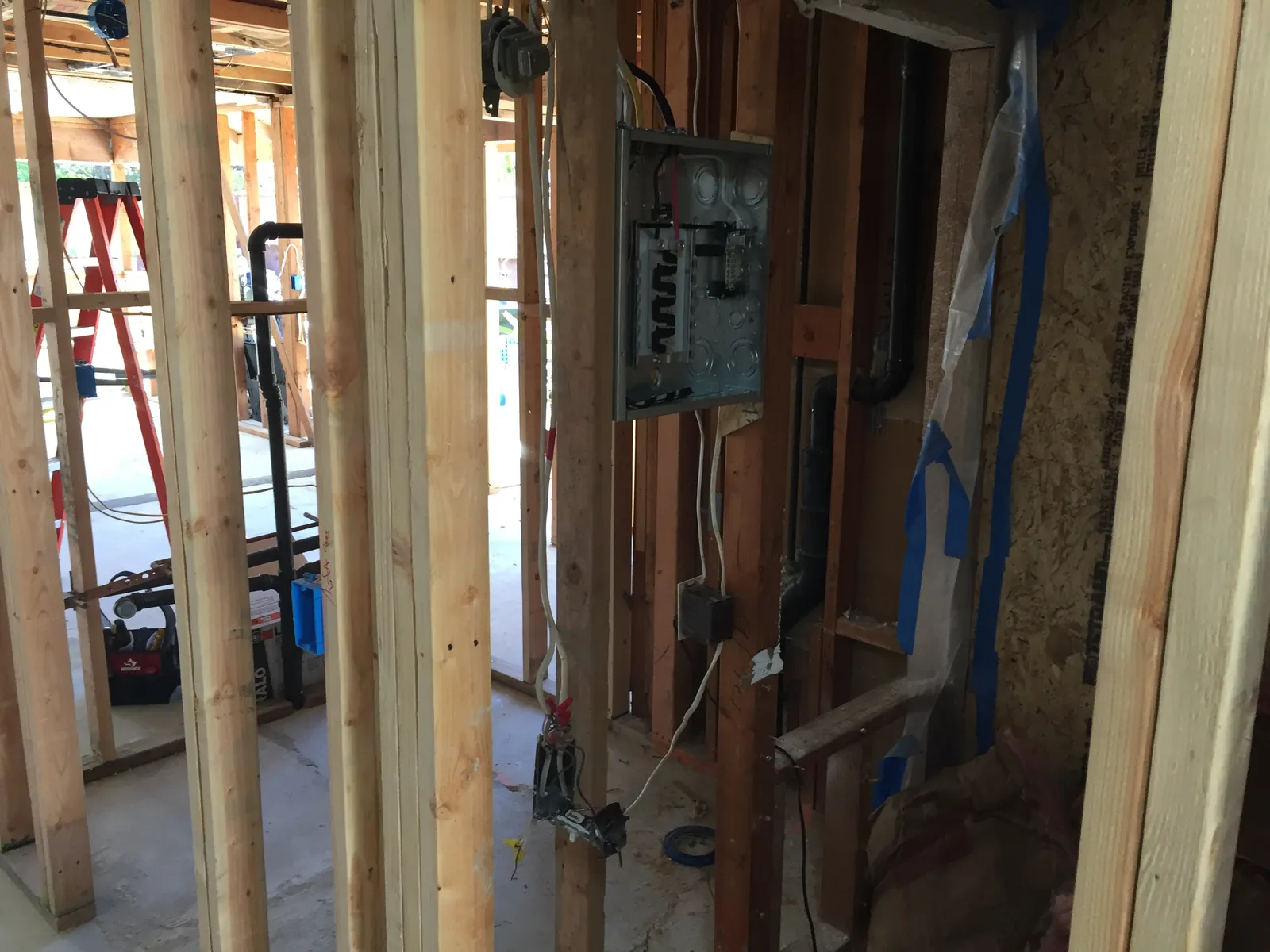 Open wood stud framing during a remodel with an electrical subpanel, rough wiring and a ladder in the background.