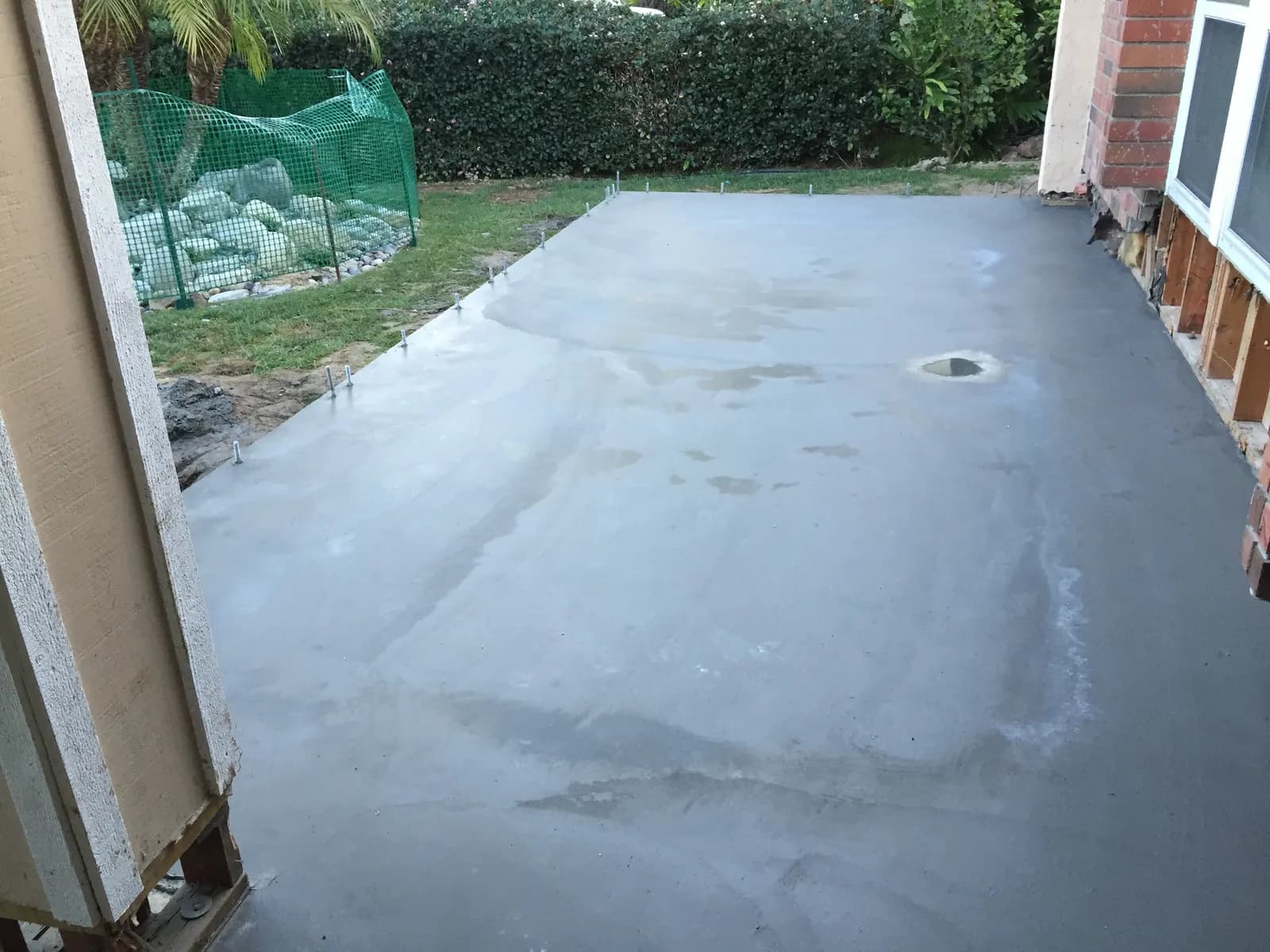A freshly poured concrete slab with anchor bolts along the edges beside a house and green backyard hedge.