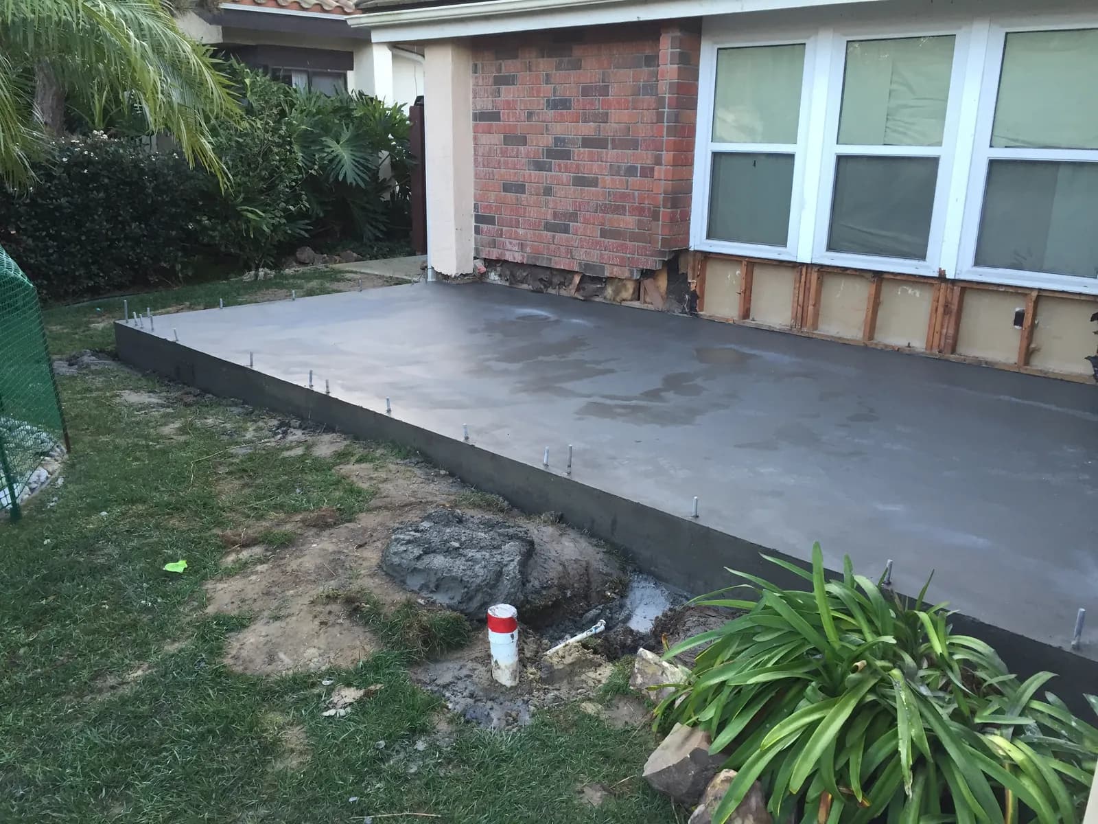 Freshly poured concrete slab with anchor bolts beside a brick house wall stripped to the framing.