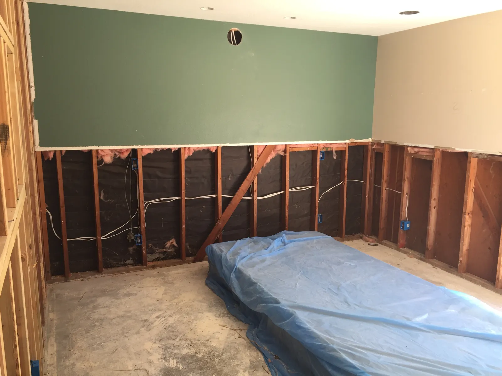 Room mid-demolition with lower drywall removed, exposed studs and wiring, and plastic sheeting over materials on the floor.