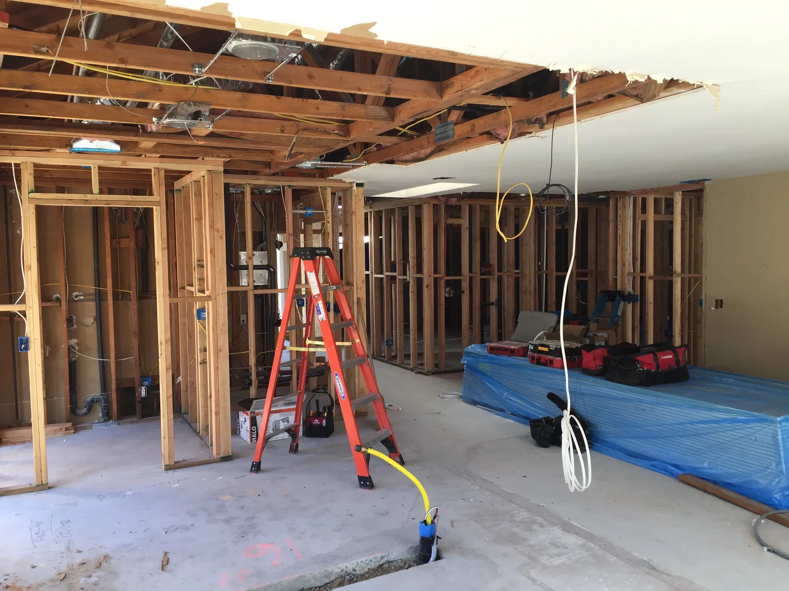 Interior remodel in progress with exposed wall framing, open ceiling joists, wiring, and an orange stepladder.