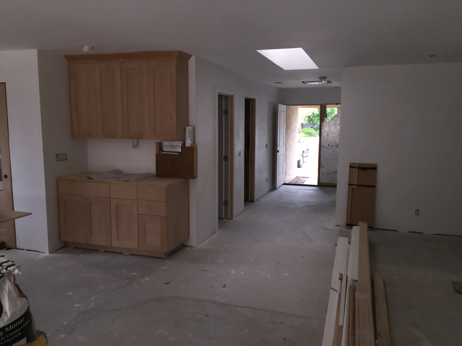 Home interior mid-remodel with new unfinished cabinets installed, bare concrete floors, and lumber stacked near the open front door.