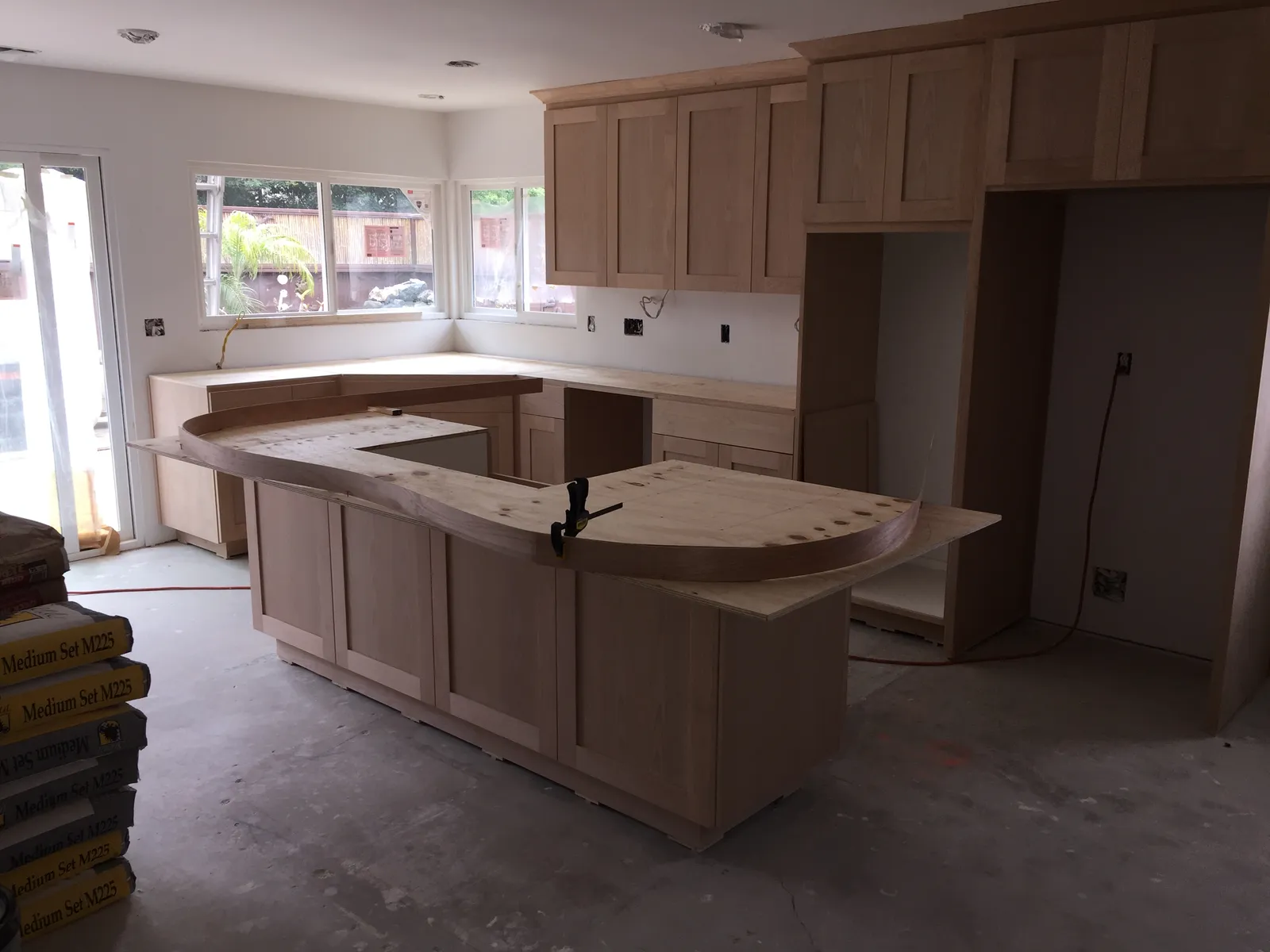A kitchen mid-remodel with unfinished cabinets installed, a curved plywood countertop template, and bags of mortar.