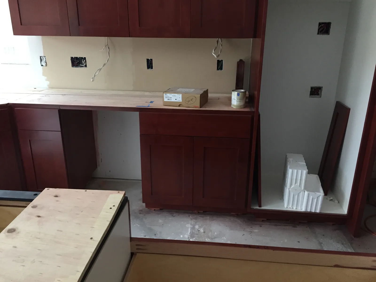 Cherry base and wall cabinets being installed in a kitchen with plywood counters and unfinished walls.